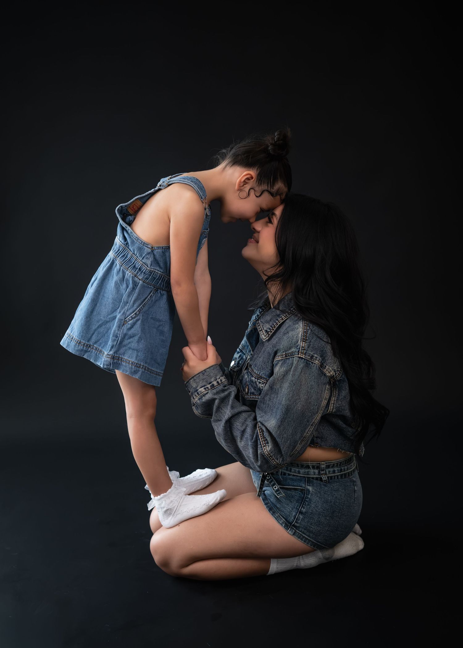 A woman in denim sits on the floor touching foreheads with a young girl in a denim dress against a dark background.