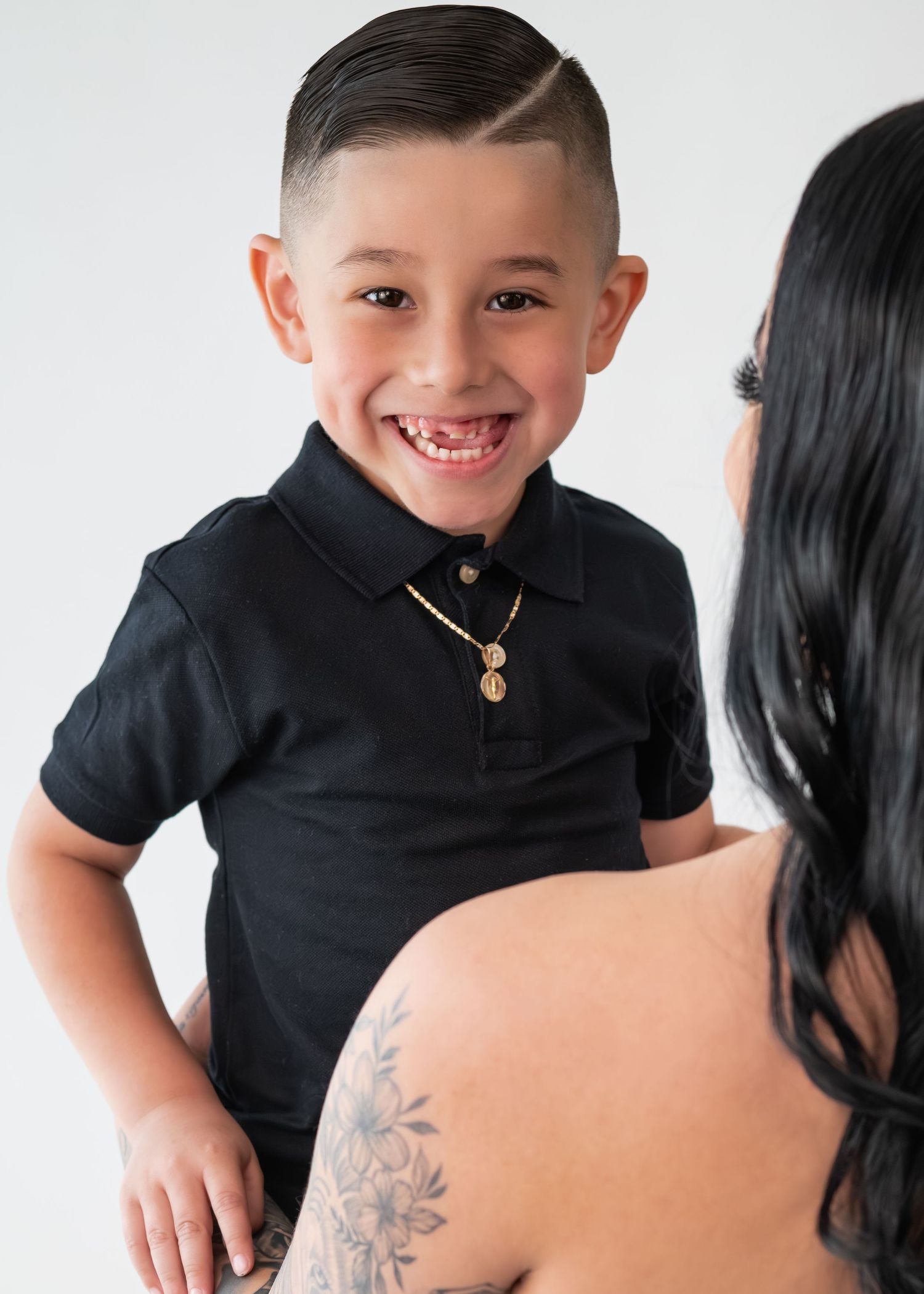 Smiling young boy in black polo with gold necklace being held by tattooed woman in studio.