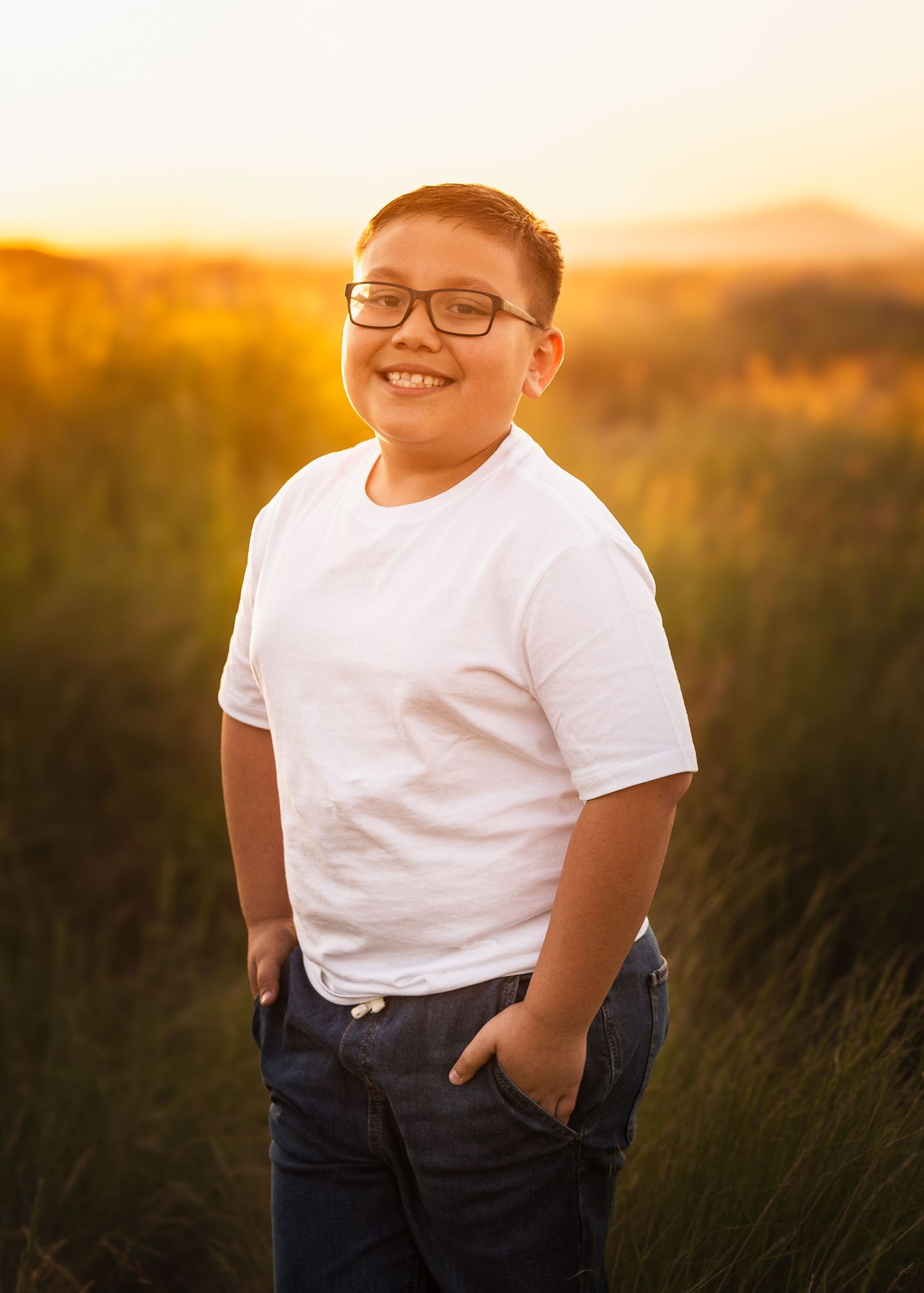 Portrait of a child in glasses and white t-shirt smiling outdoors during golden hour sunset with tall grass in background.
