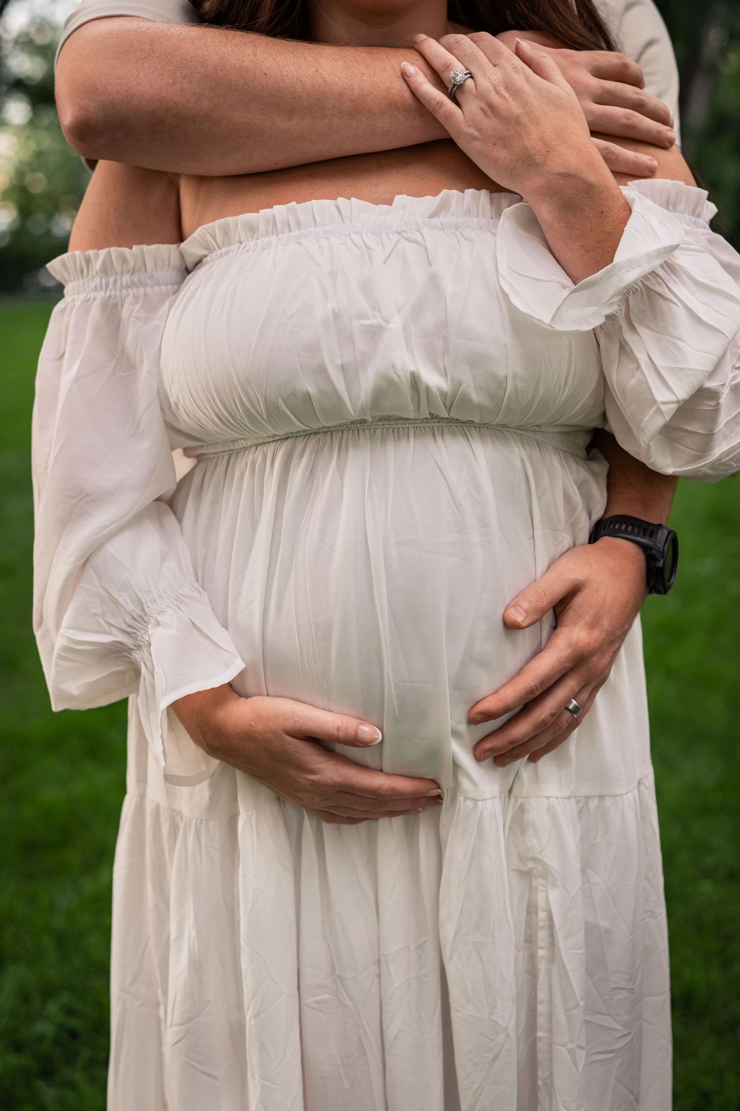 Close-up of pregnant belly in white off-shoulder maternity dress with hands tenderly cradling the bump outdoors.