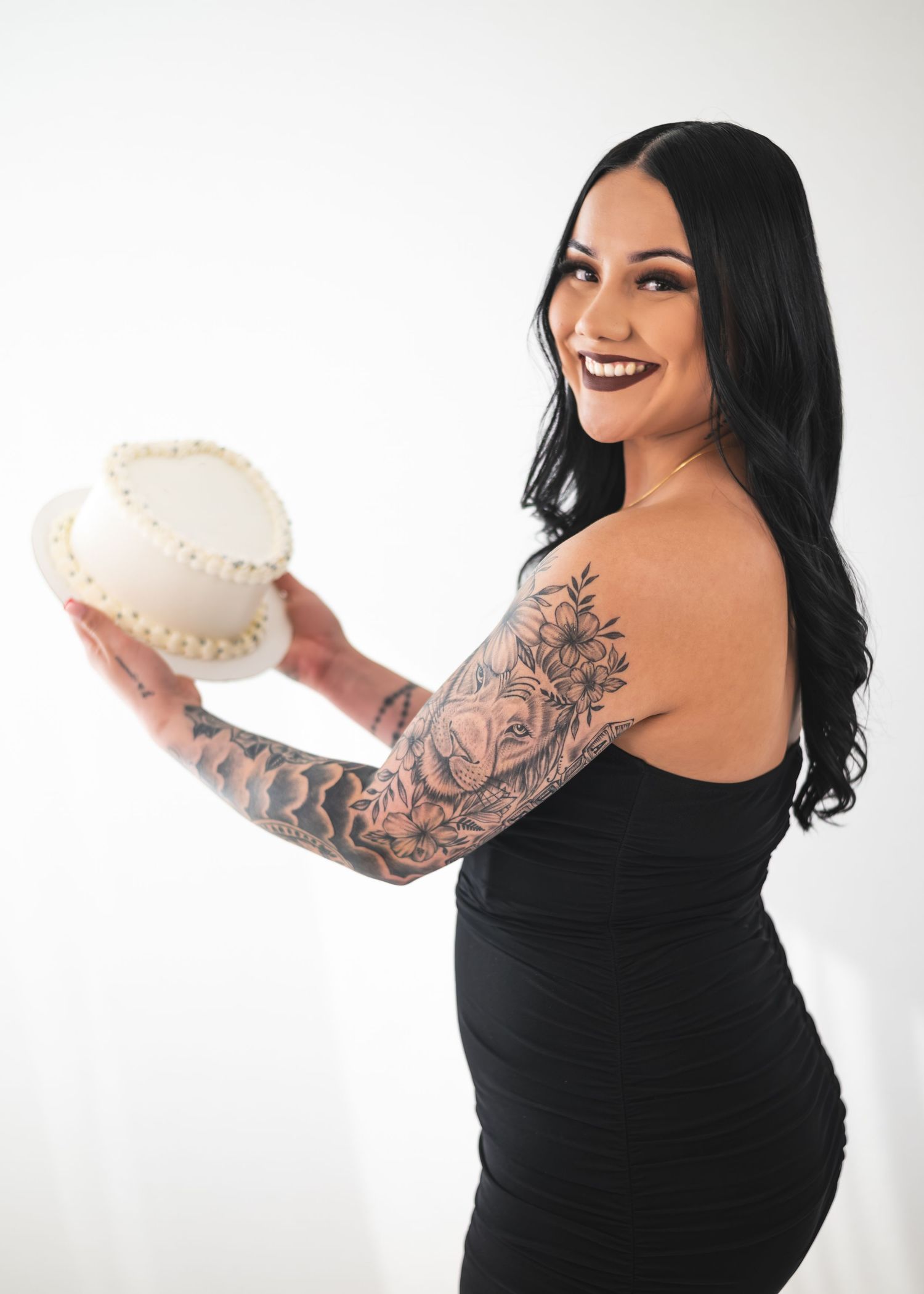 Tattooed woman in black dress smiling and holding a white cake, showcasing elegant tattoo sleeve art.