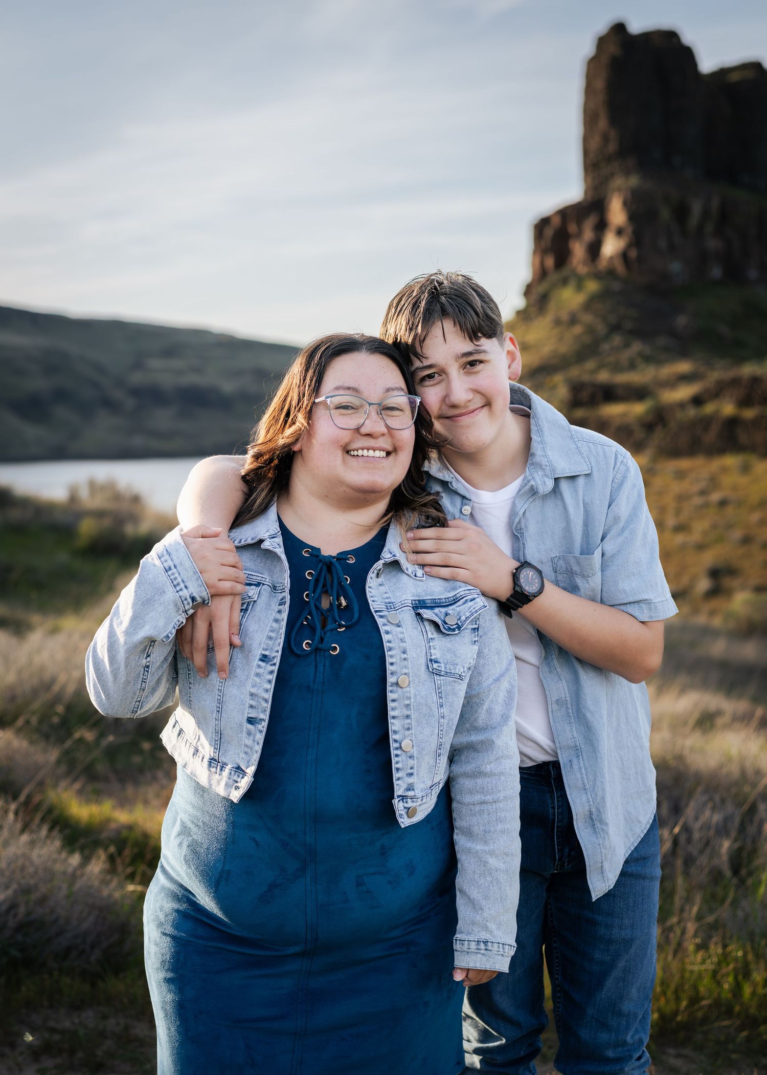 Two smiling people in denim outfits pose together outdoors near a rocky hillside and lake at golden hour.