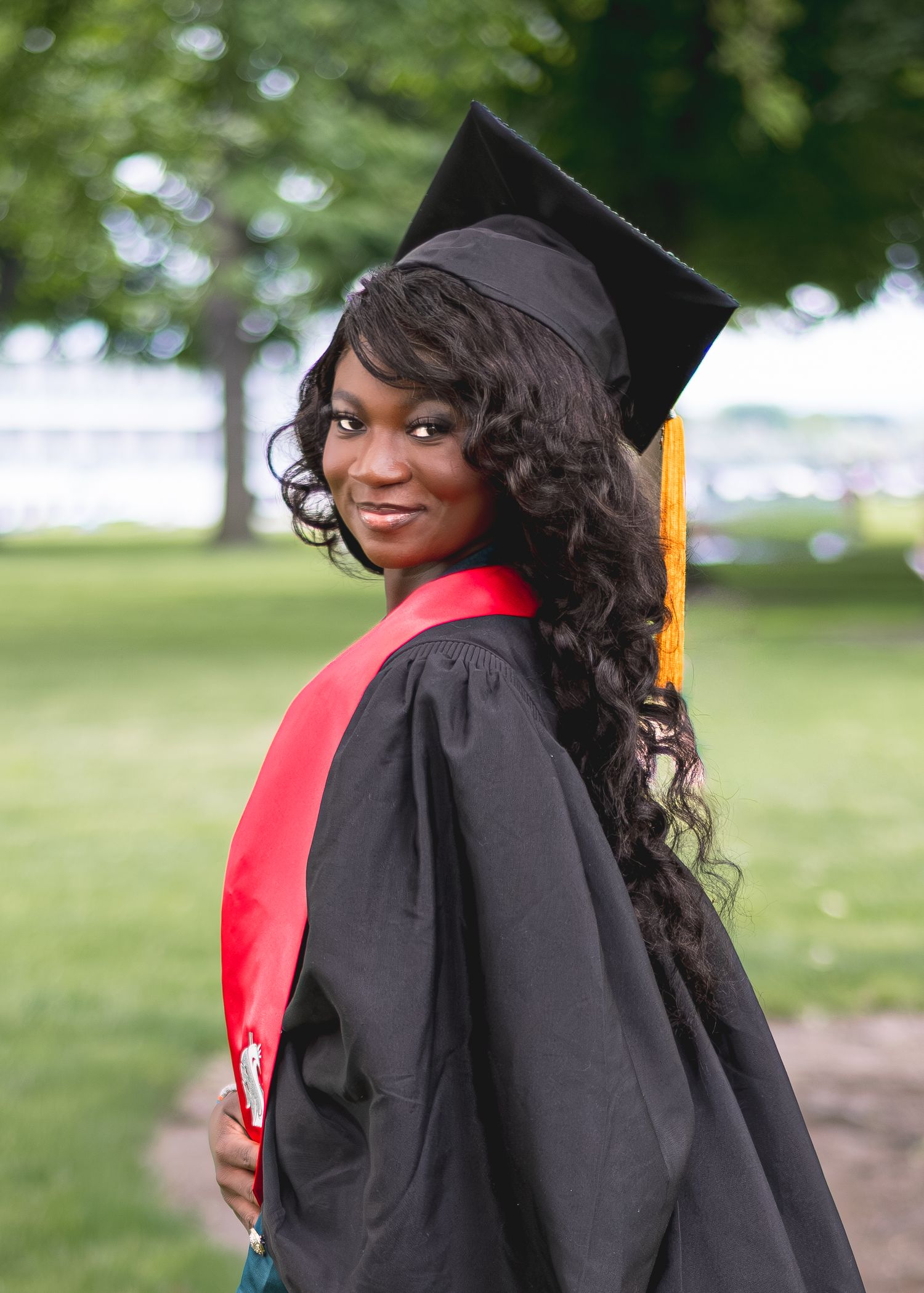 Young woman in black graduation cap and gown with red sash smiling outdoors on a green campus lawn.