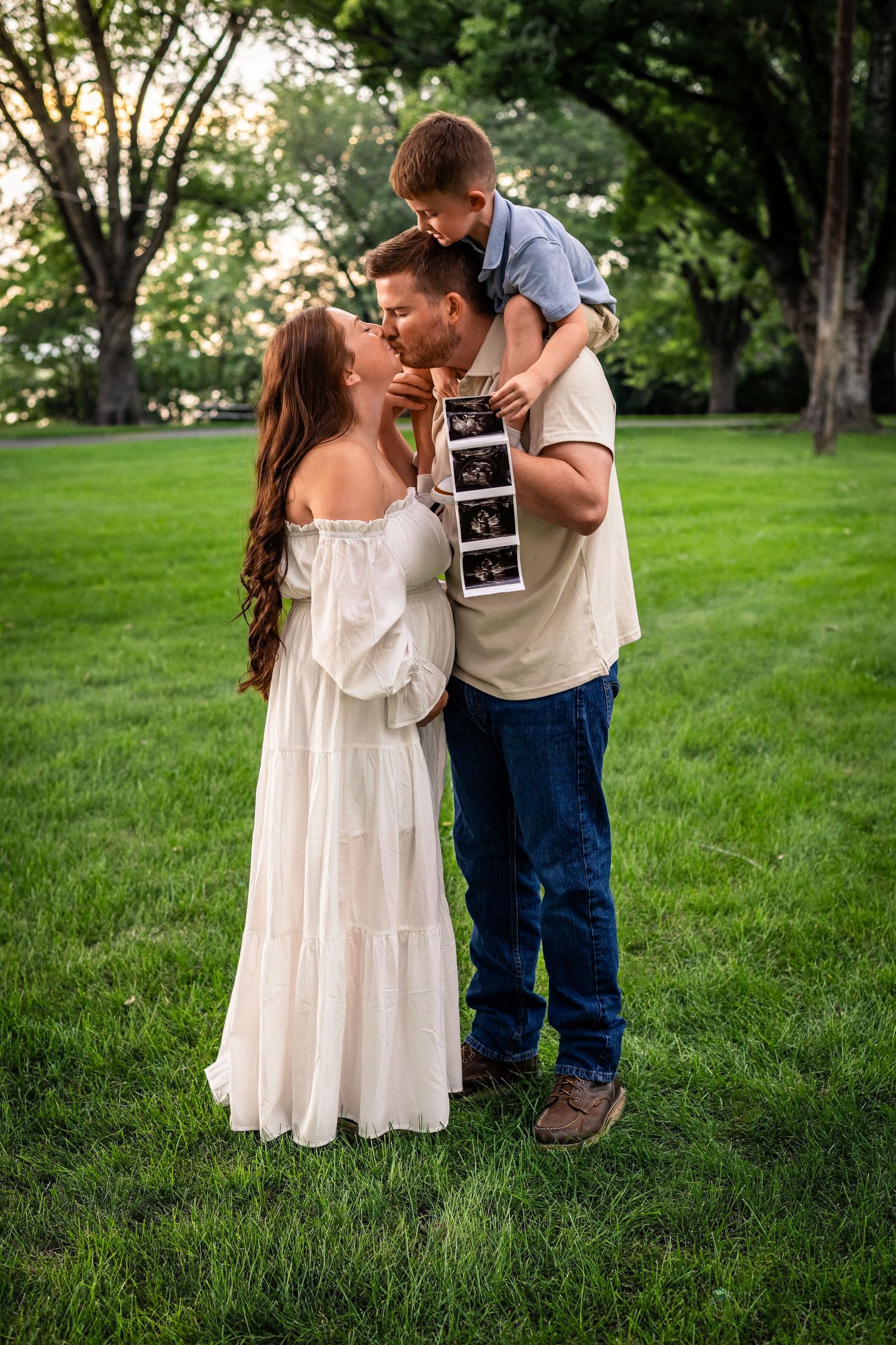 A couple in a grassy park shares a tender moment while holding an ultrasound photo during a pregnancy announcement shoot.