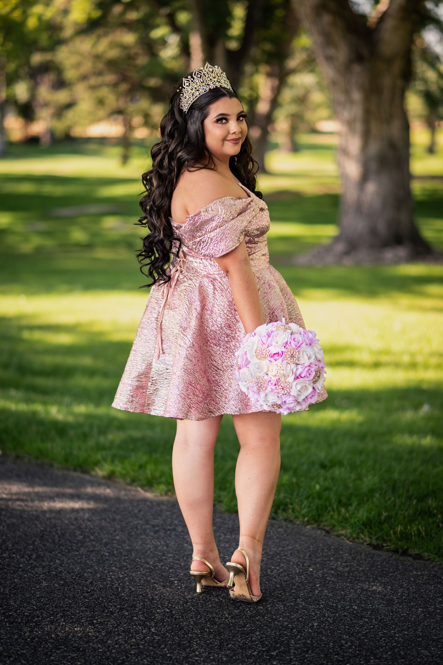 A person in a pink off-shoulder dress poses outdoors in a park setting with blurred trees in the background.