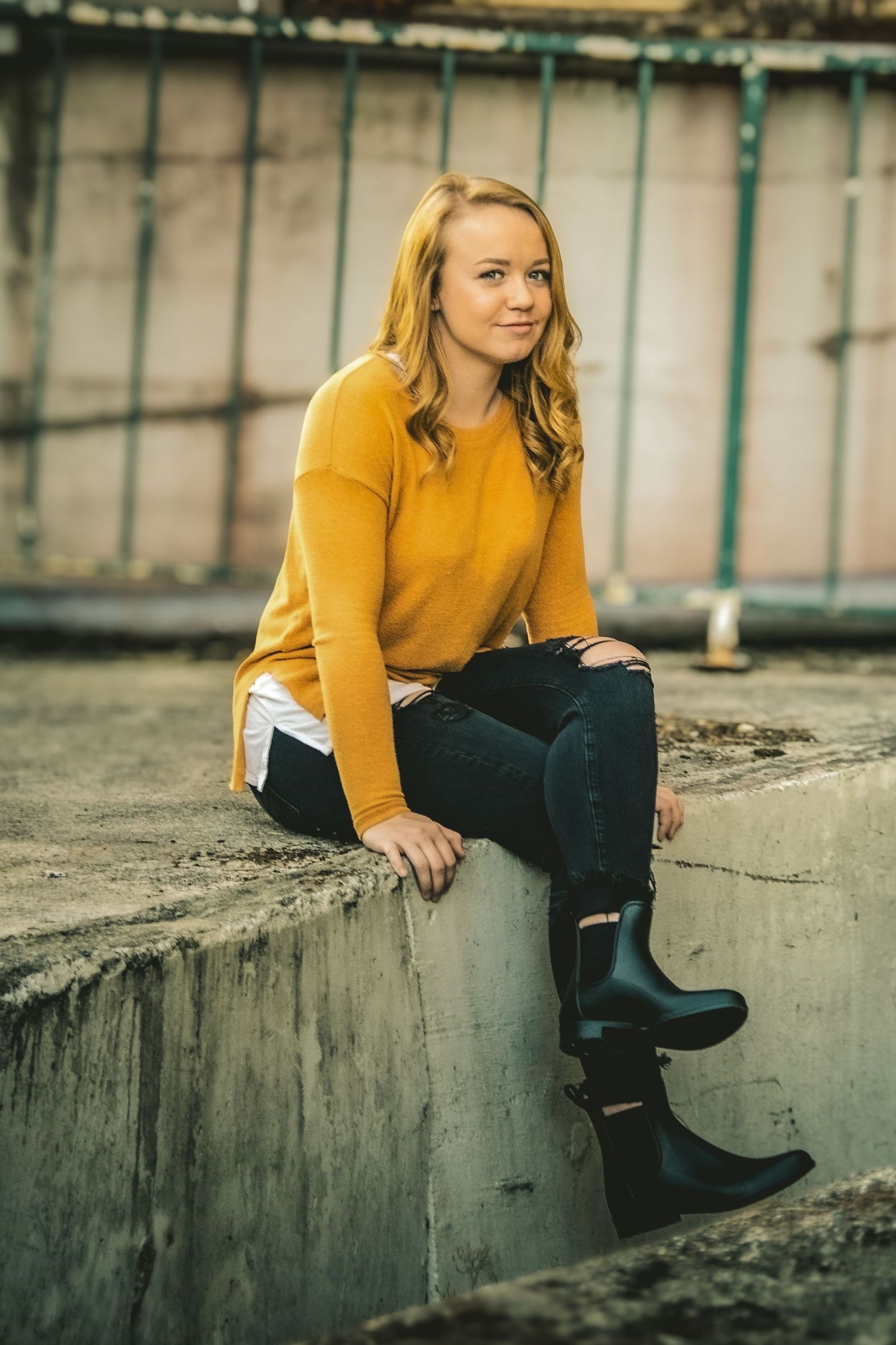 A person in a mustard yellow sweater sits casually on a concrete ledge wearing dark jeans and boots.