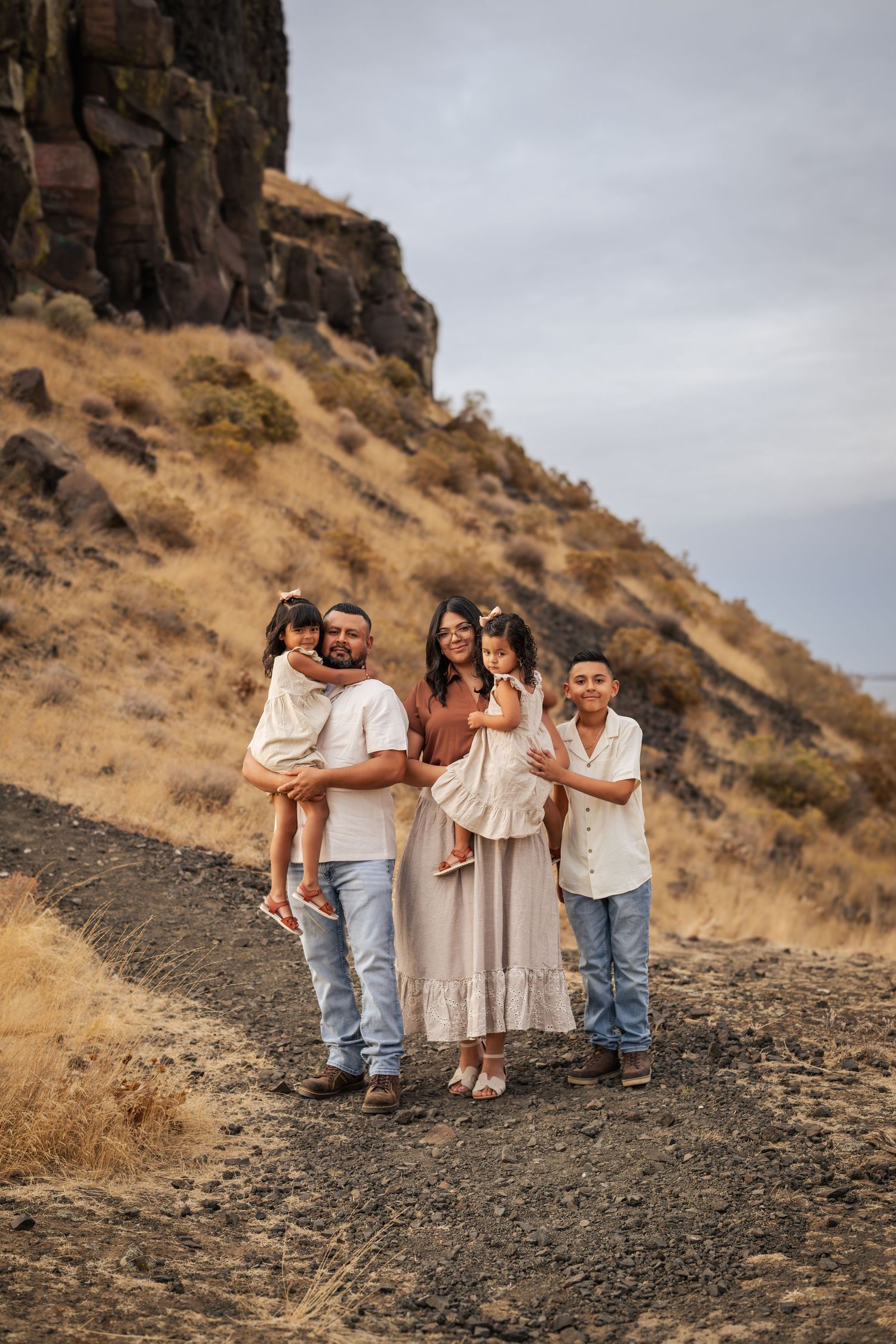 Family in coordinating neutral outfits poses together against dramatic rocky hillside at sunset.