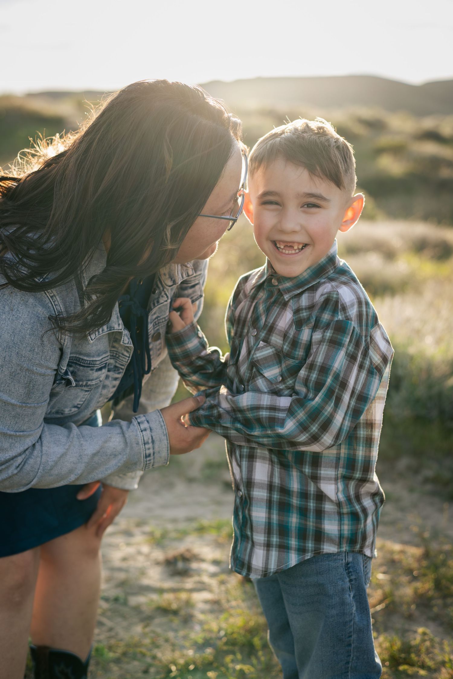 A young boy in a plaid shirt smiles joyfully while a woman with glasses leans toward him outdoors.
