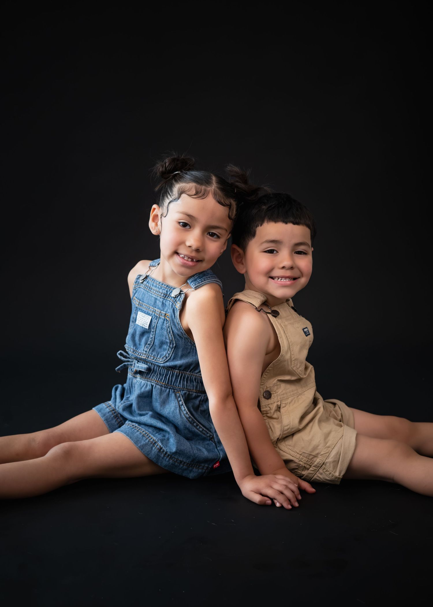 Two smiling young children sit back-to-back wearing denim and khaki rompers against a dark studio background.