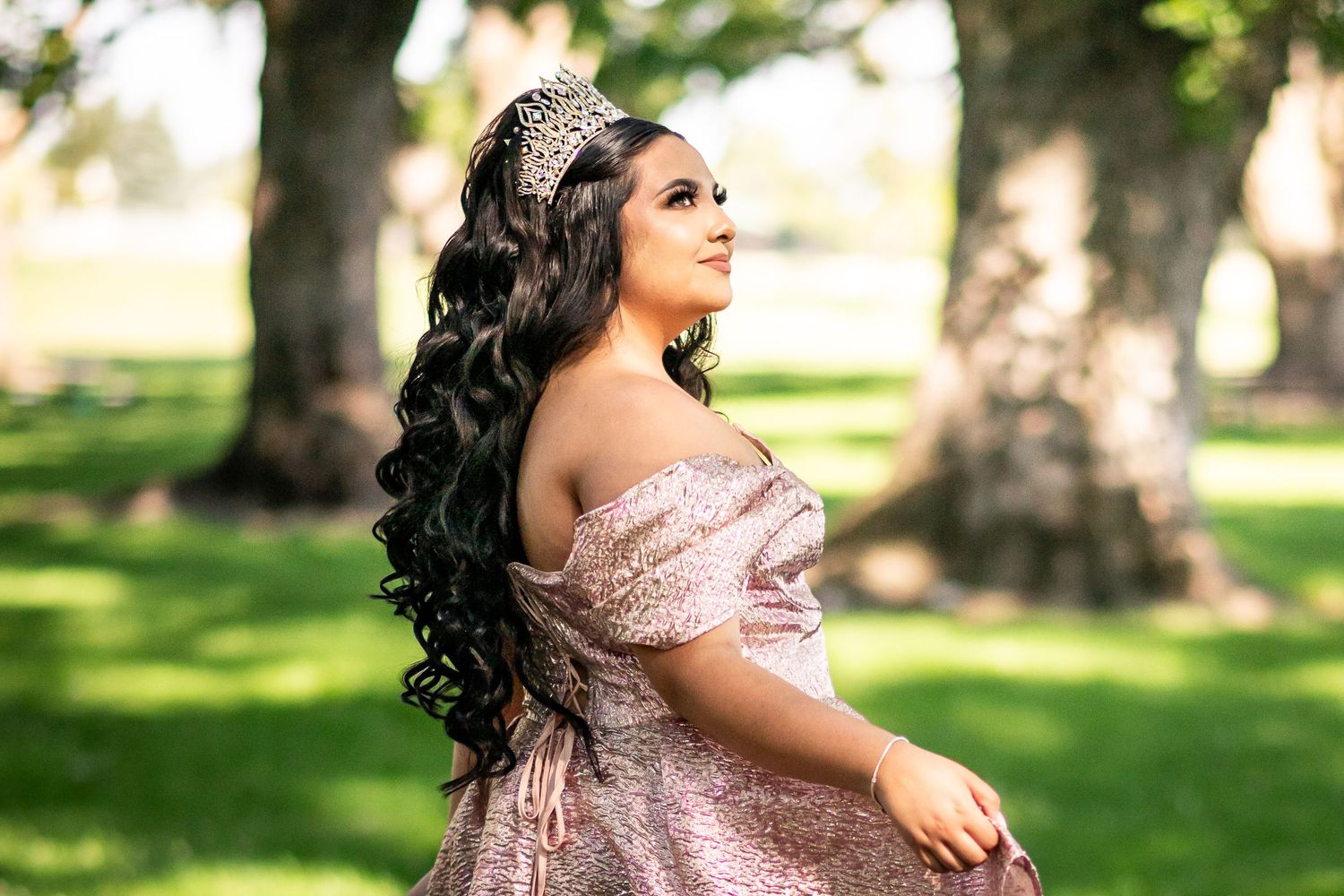 Young woman in pink off-shoulder gown and tiara with long curly hair poses outdoors among green trees.