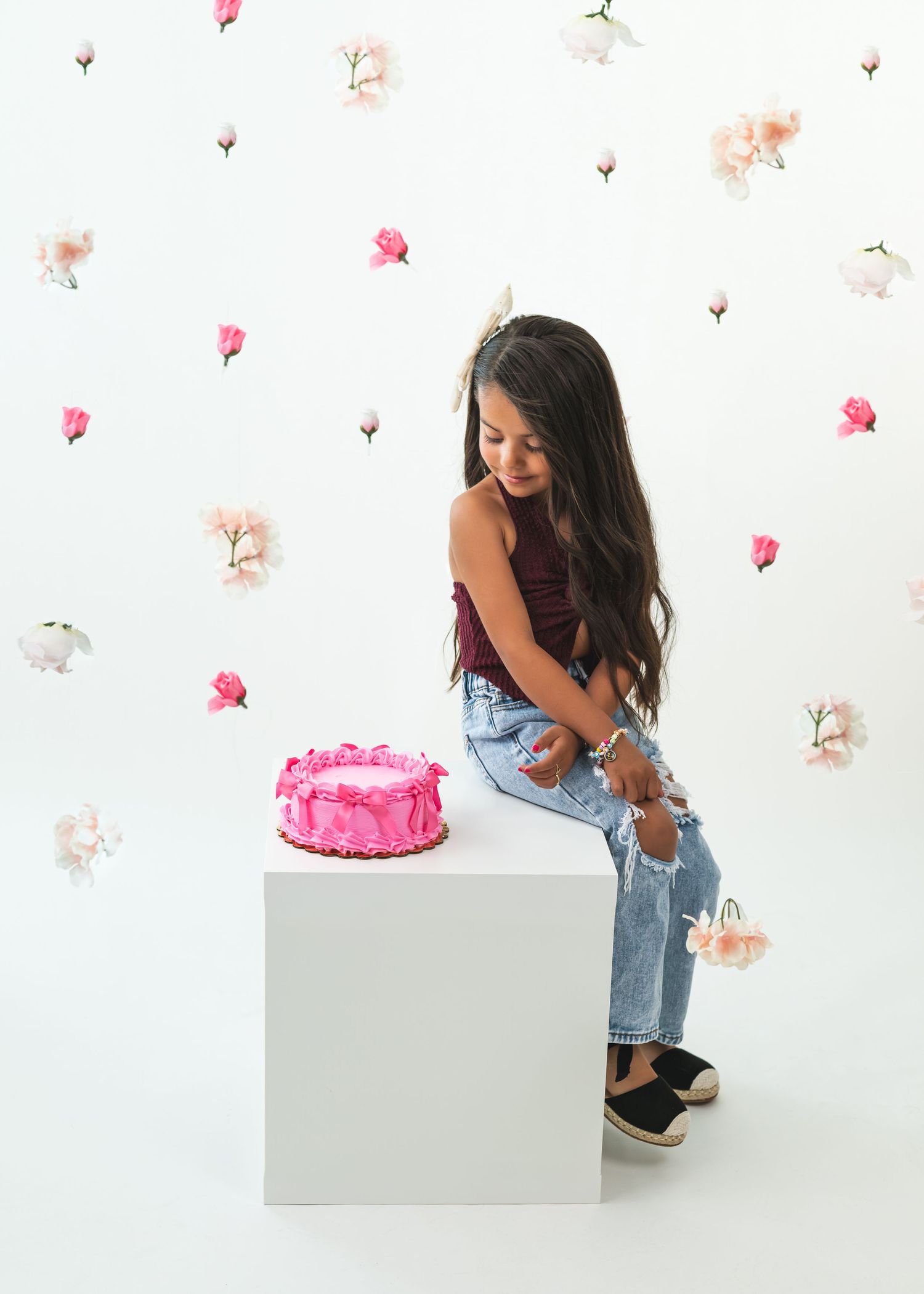 Young girl in jeans sits on white cube with pink birthday cake, floral wallpaper background.