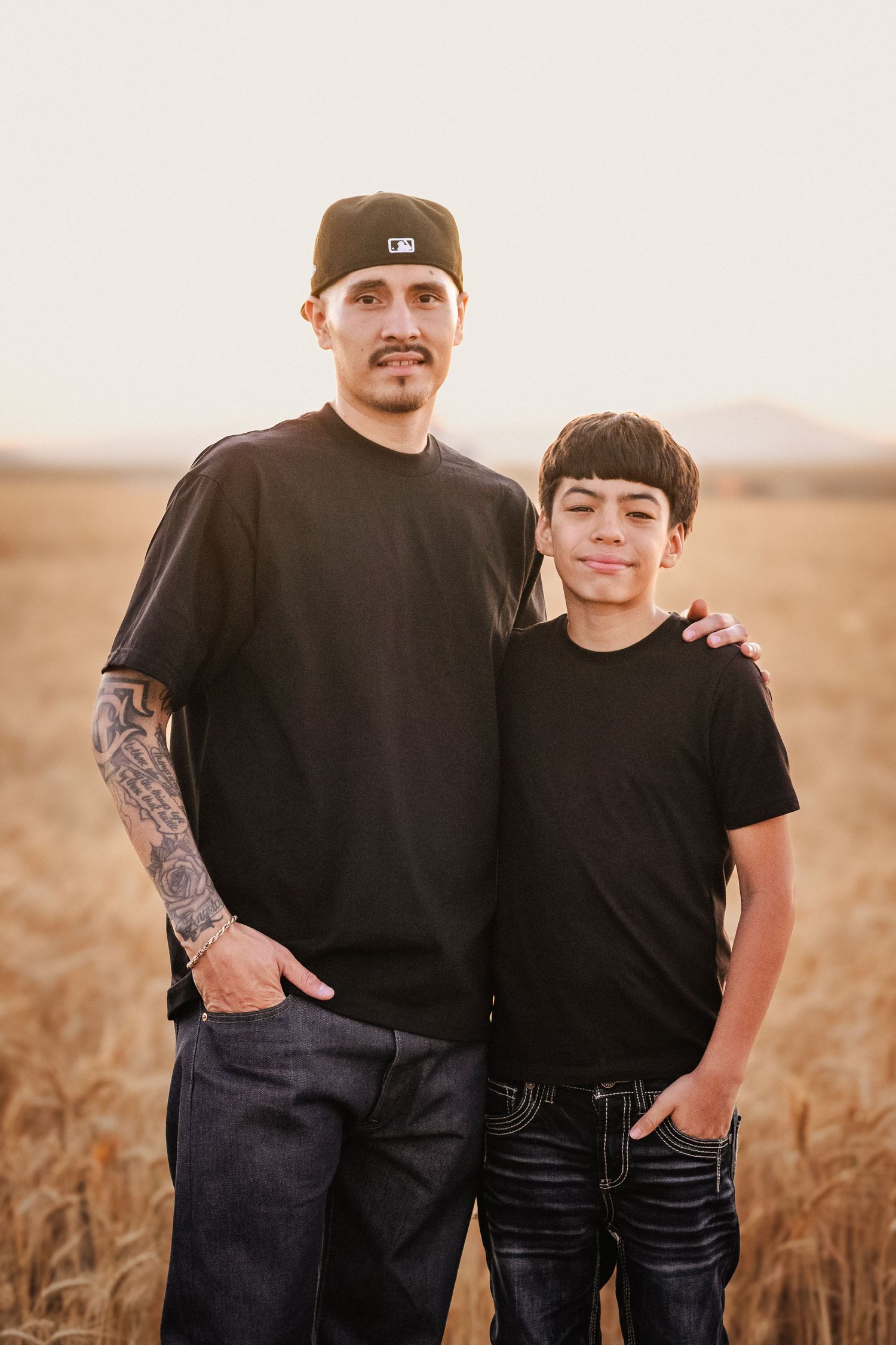 Man with tattoos and cap poses with young boy in black shirts in golden wheat field at sunset.