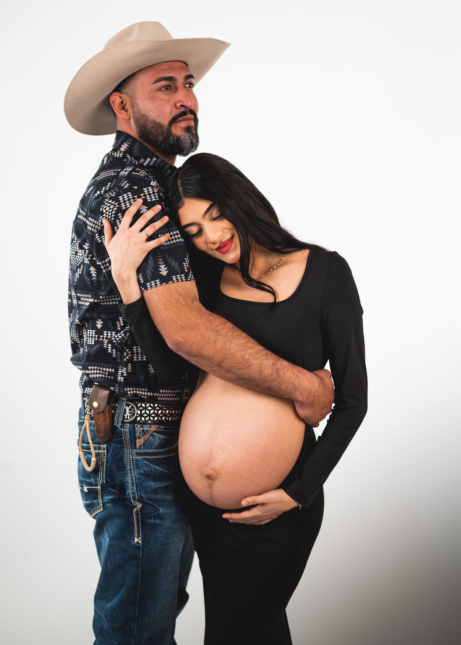 Couple maternity photo with man in cowboy hat embracing pregnant woman in black outfit against white background.