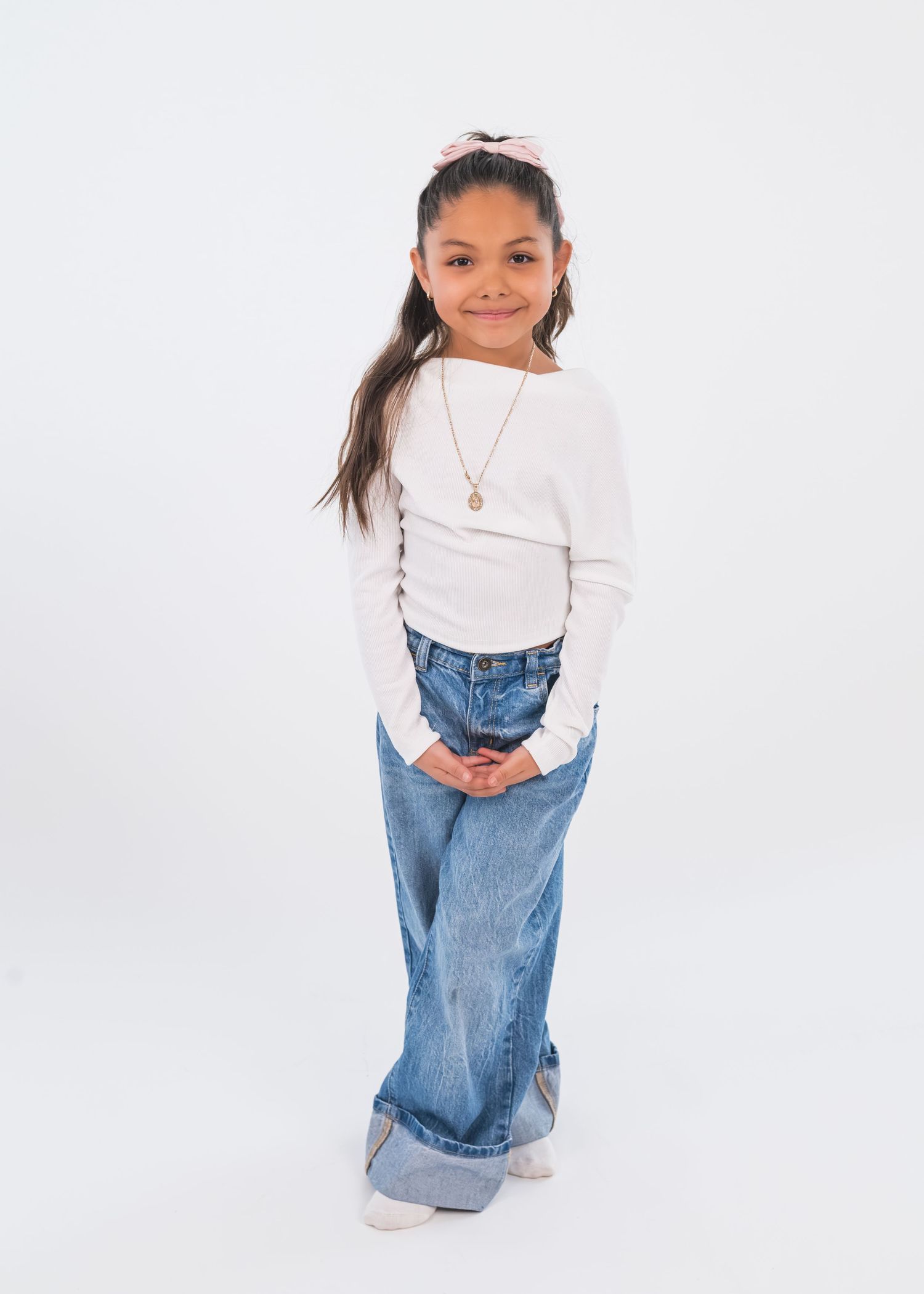 Young girl wearing a white long-sleeve top, wide-leg jeans, and a pink bow headband, posing against a white background.