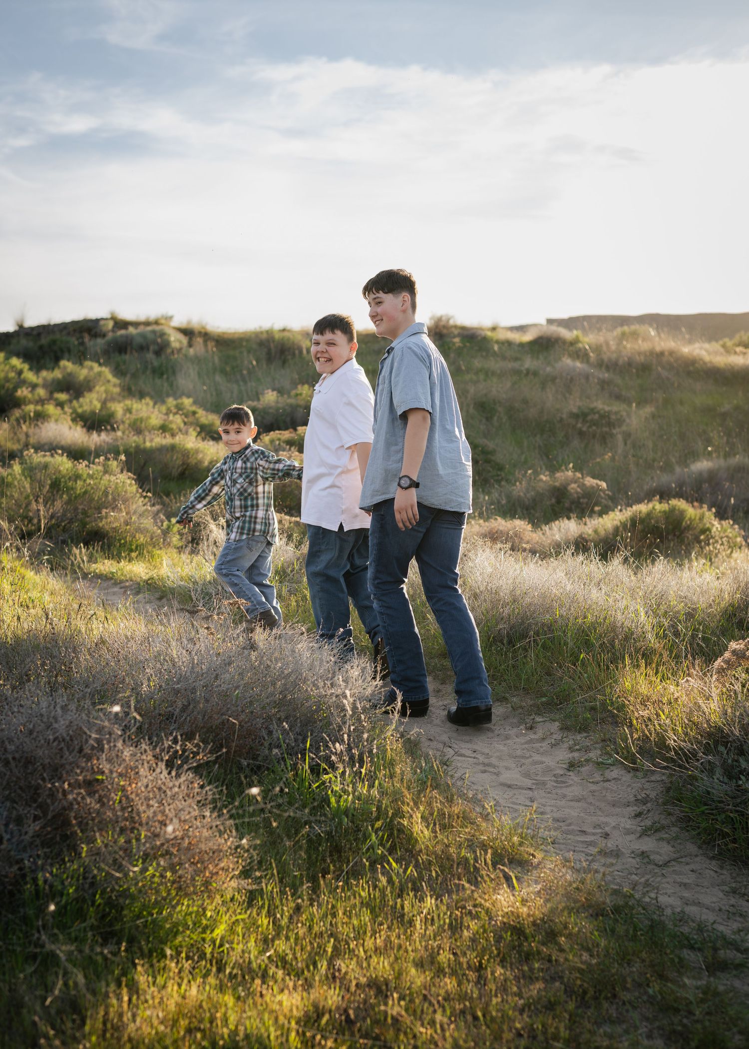 Three boys walking on a dirt trail through green hills under a cloudy sky.