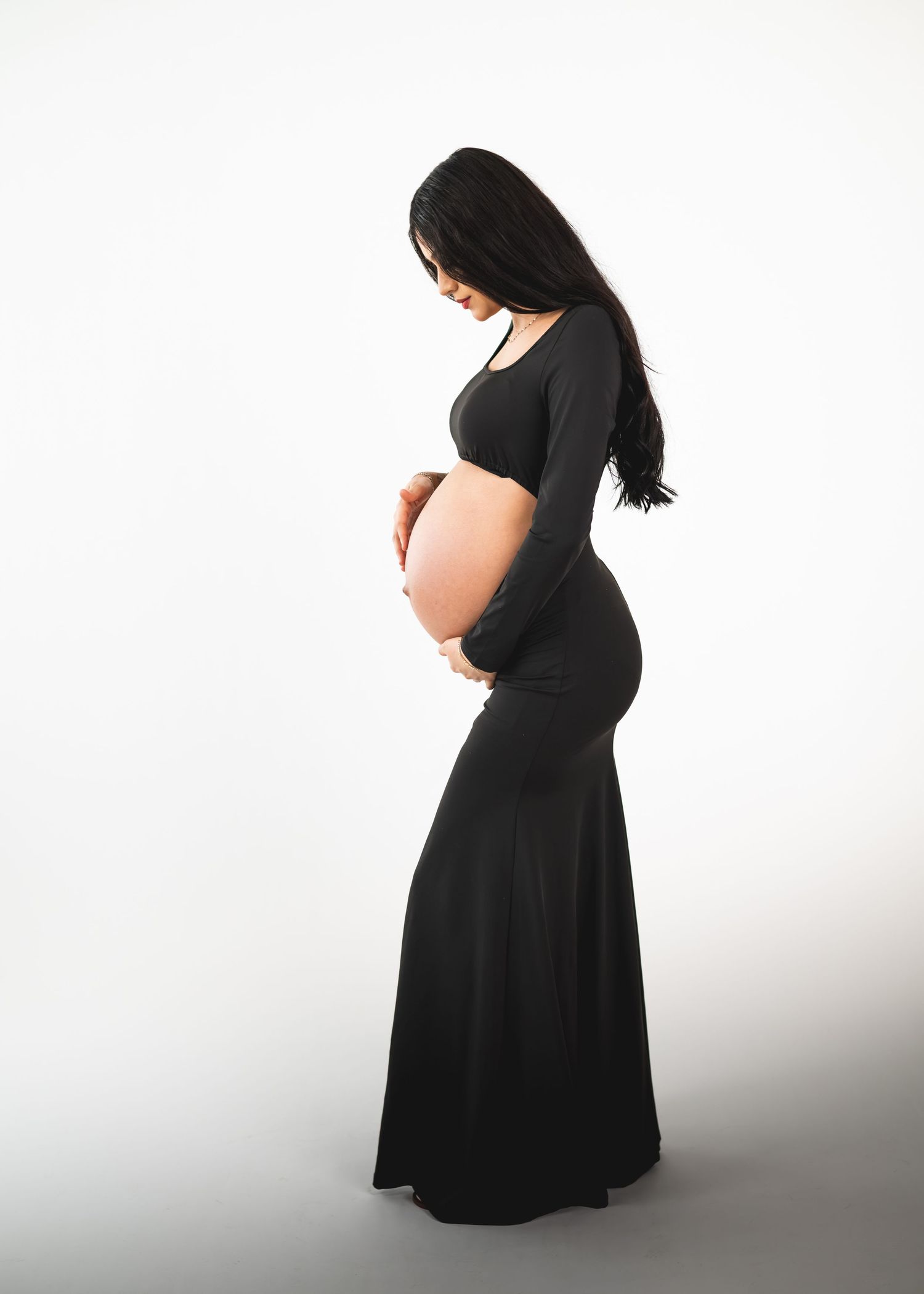Pregnant woman in black maternity gown cradling baby bump, side profile studio photo on white background.