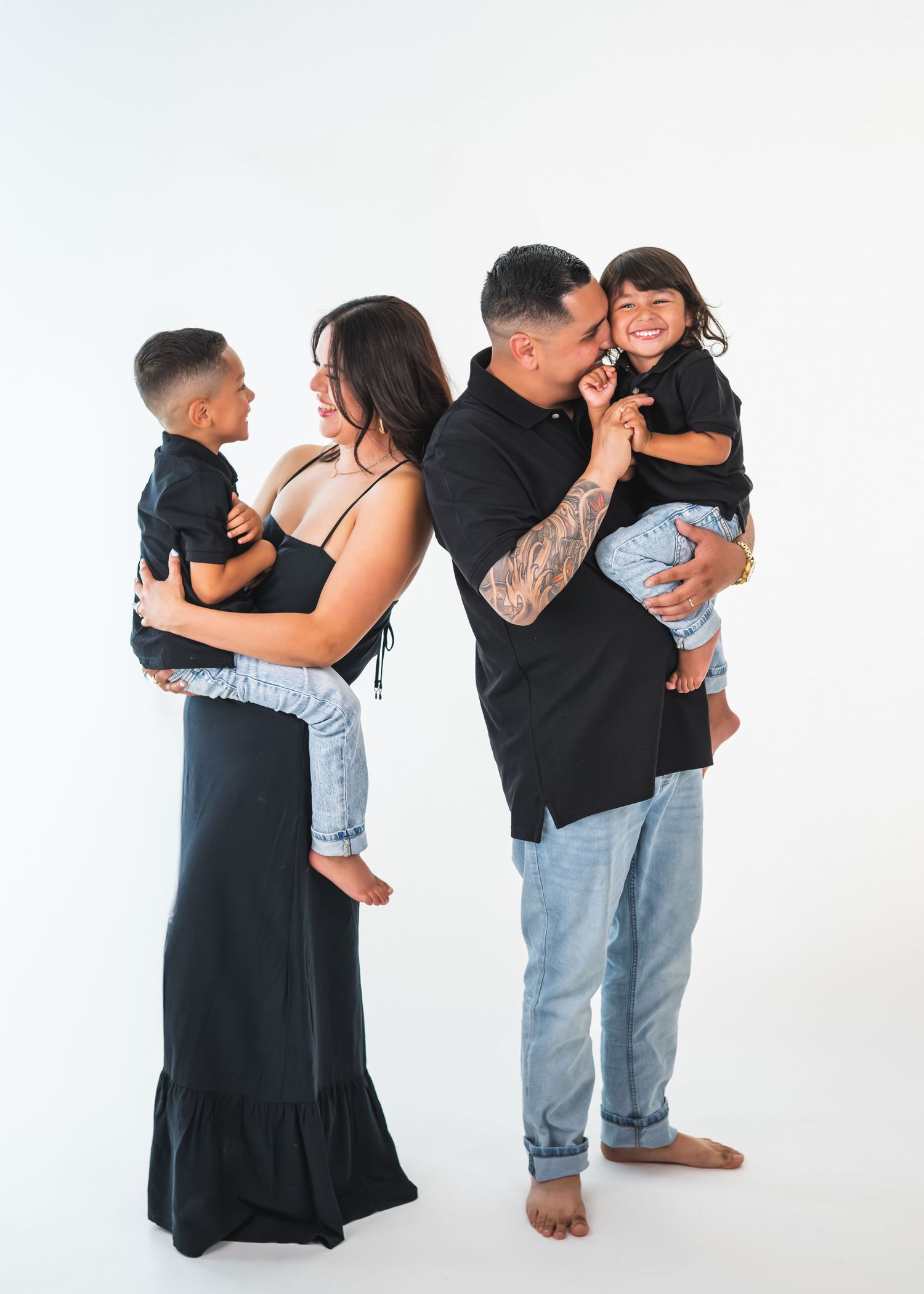 Family of four in matching black outfits poses on white background, parents holding young children, smiling joyfully.
