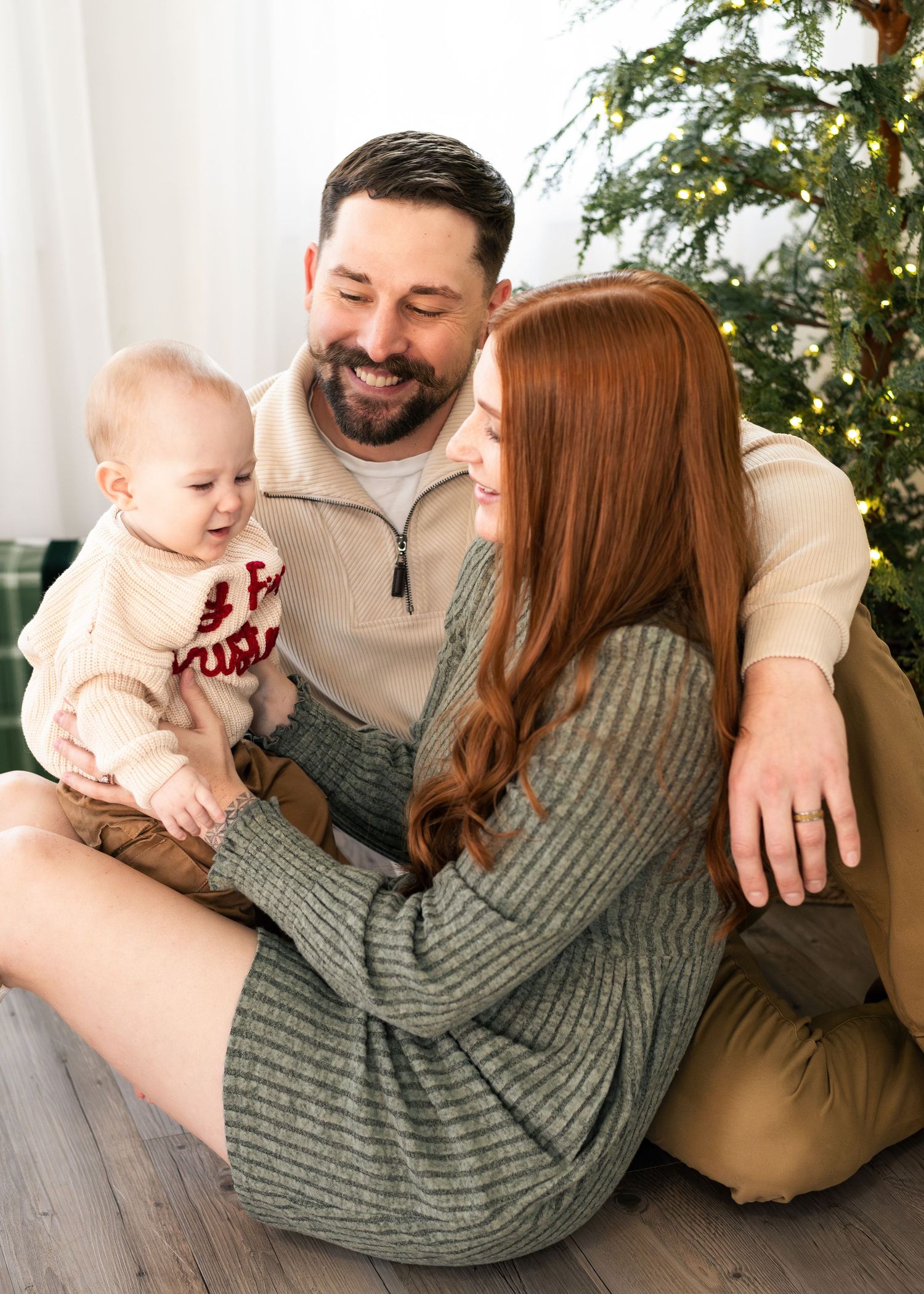 Family of three sharing tender moments on floor by Christmas tree in casual winter attire.
