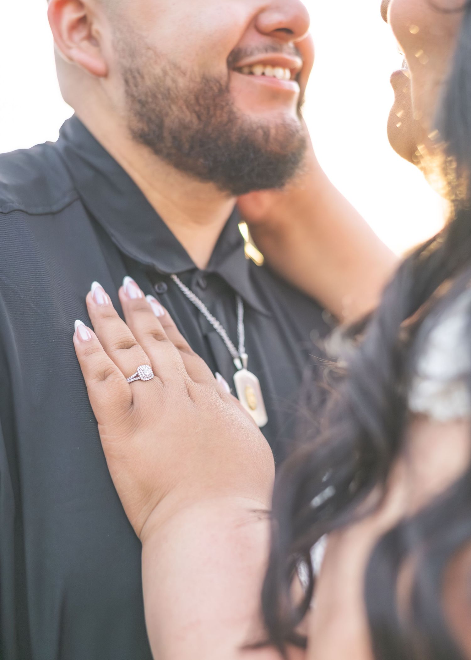Close-up of a woman's hand with an engagement ring resting on a smiling man's chest during a romantic outdoor photo session.