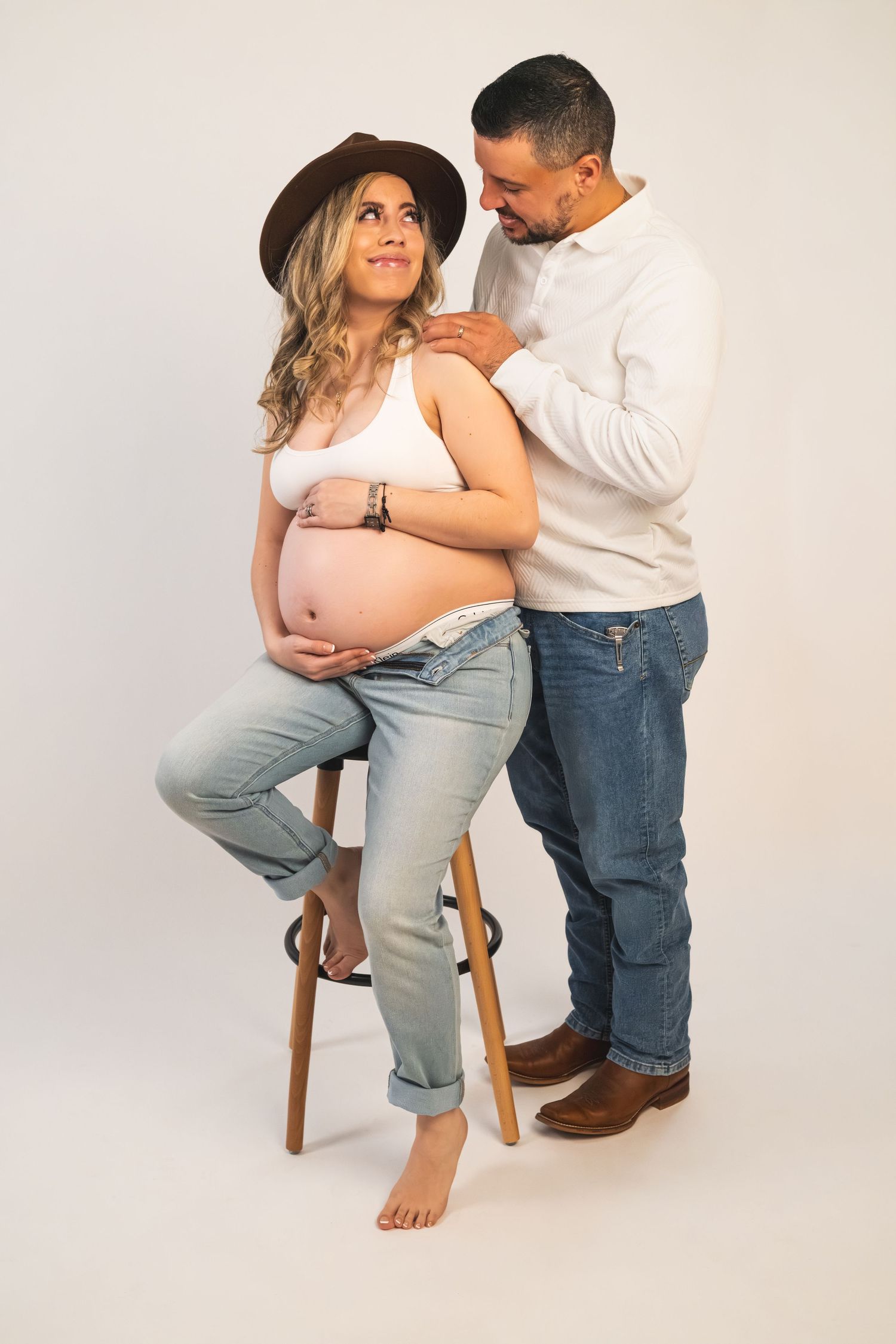 A couple shares an intimate moment in a minimalist studio setting with white background and wooden stool.