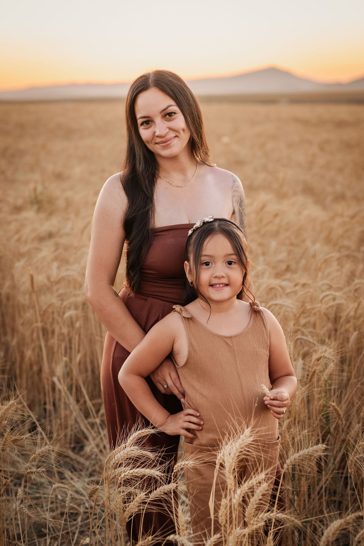 A woman and young girl smile together in a golden wheat field at sunset, wearing warm-toned dresses.