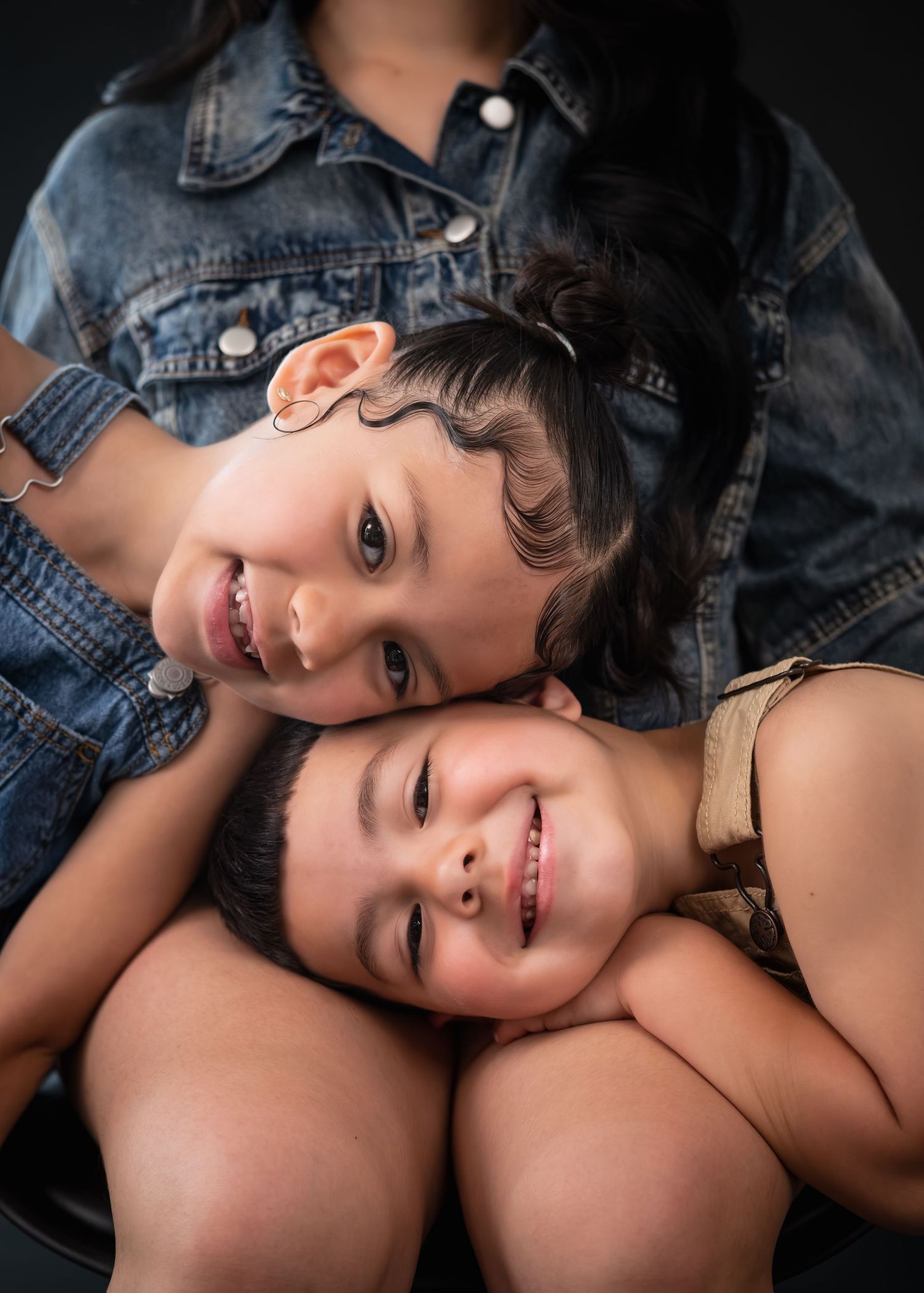 Two smiling children cuddle together in denim outfits against a dark studio background.
