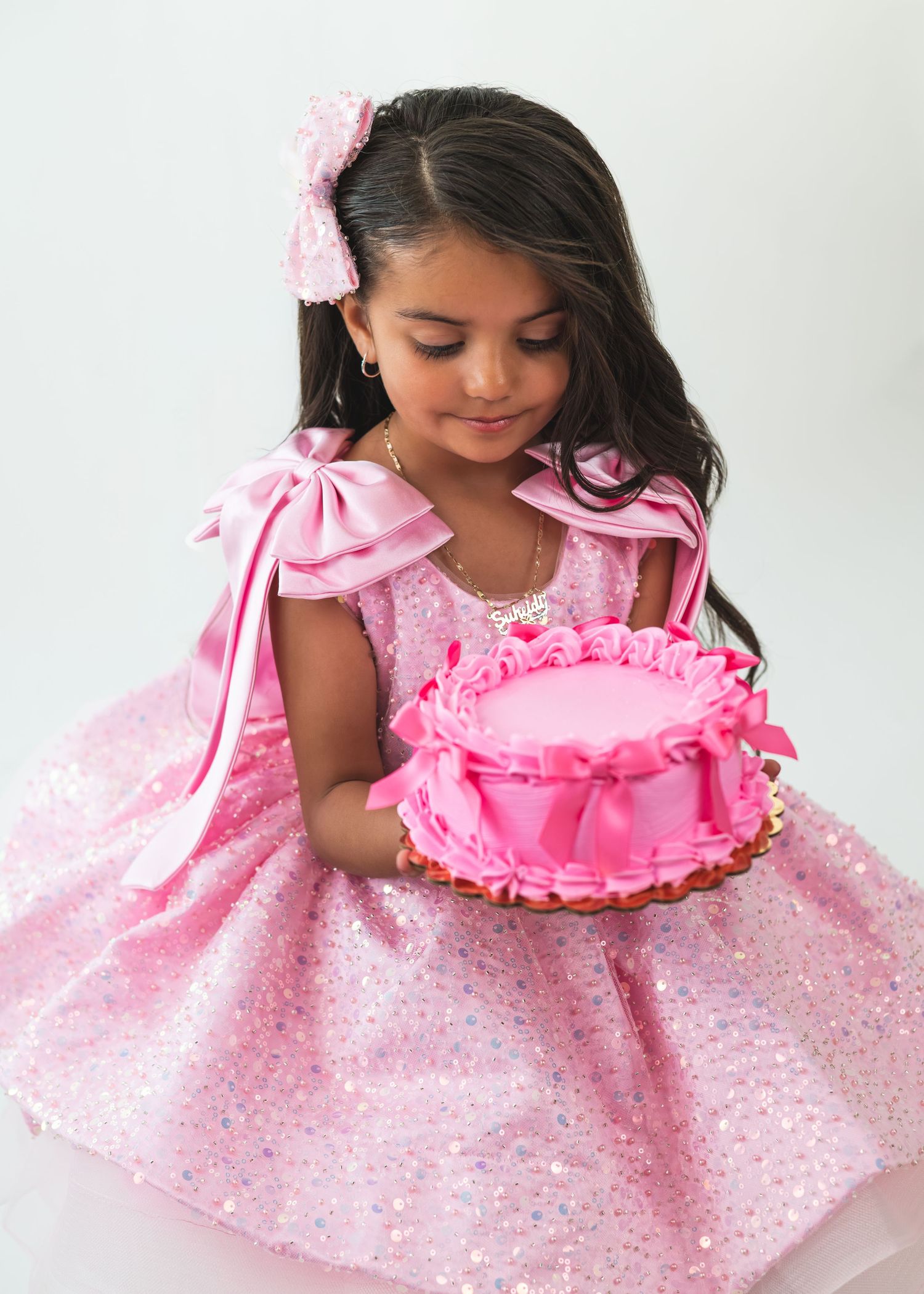Young girl in pink sequin dress holding a pink birthday cake, wearing a bow hair accessory.