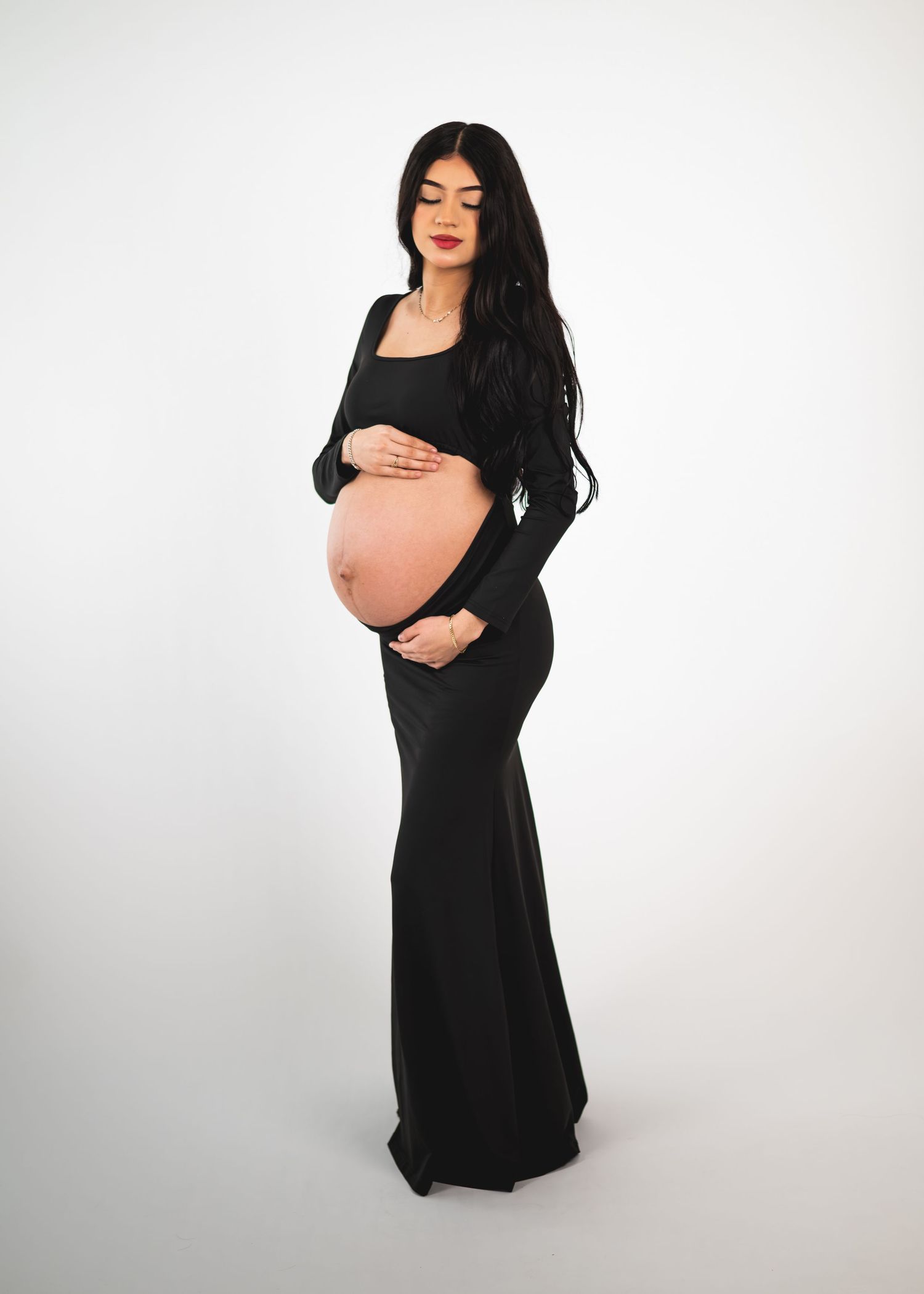 Pregnant woman in black maternity gown cradling baby bump, long dark hair, white background.