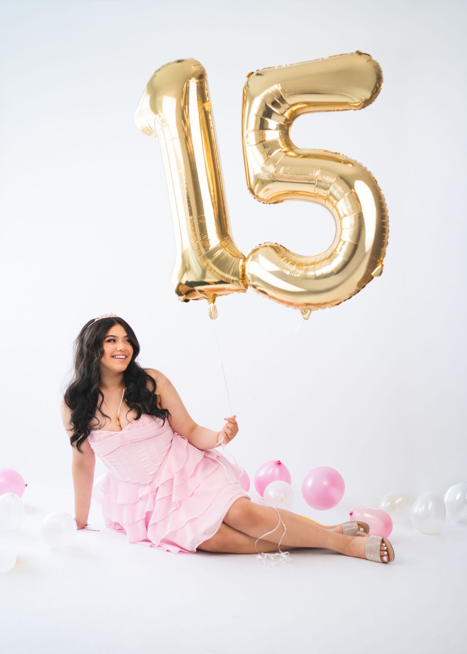 Young woman in pink dress sitting on floor holding gold number 15 balloons, celebrating a quinceañera birthday.