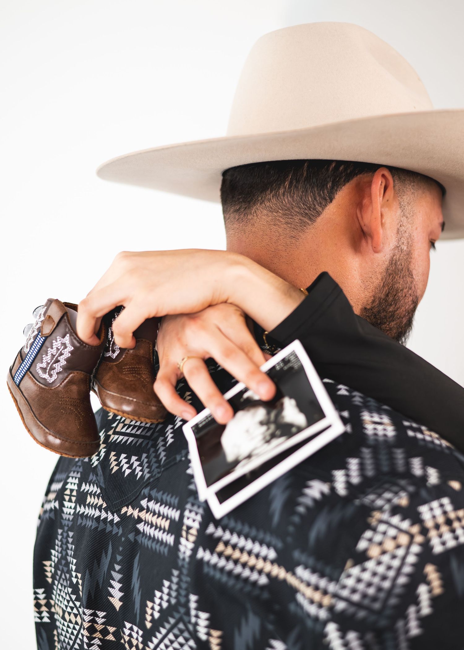 Man in cowboy hat holding baby boots and ultrasound photo over his shoulder, announcing pregnancy.