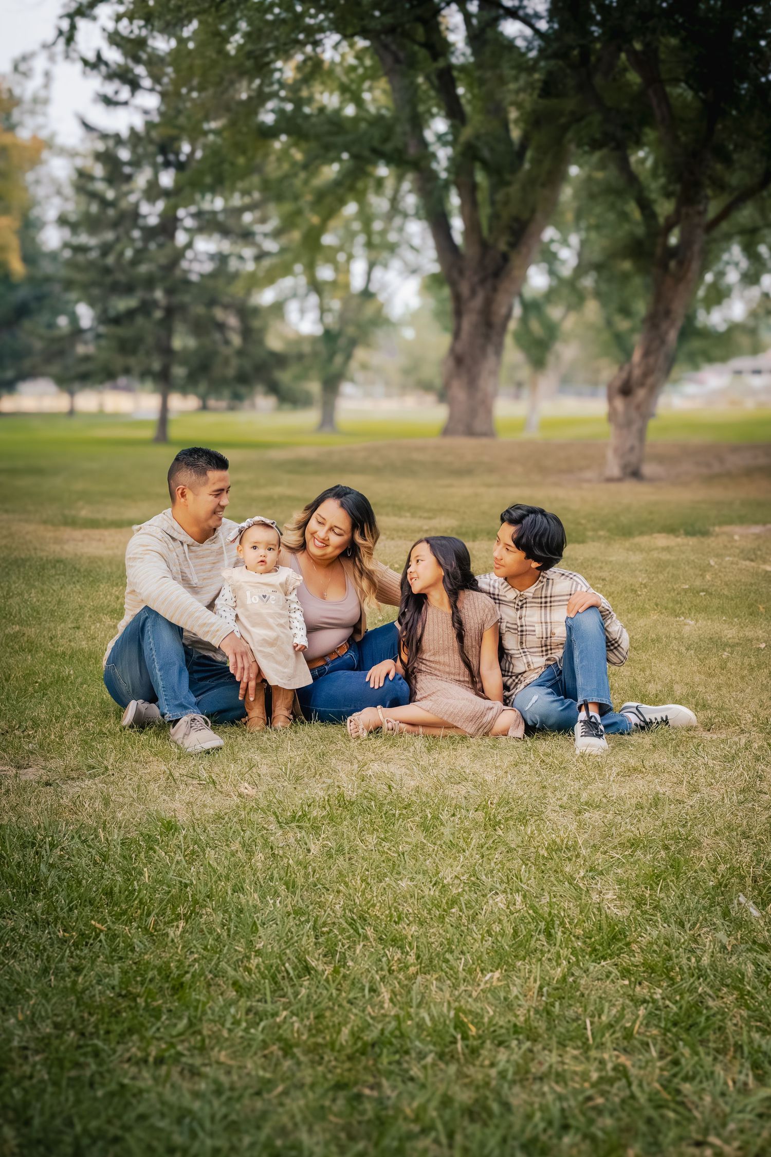 A family sits together on green grass in a park with tall trees in the background during a sunny day.
