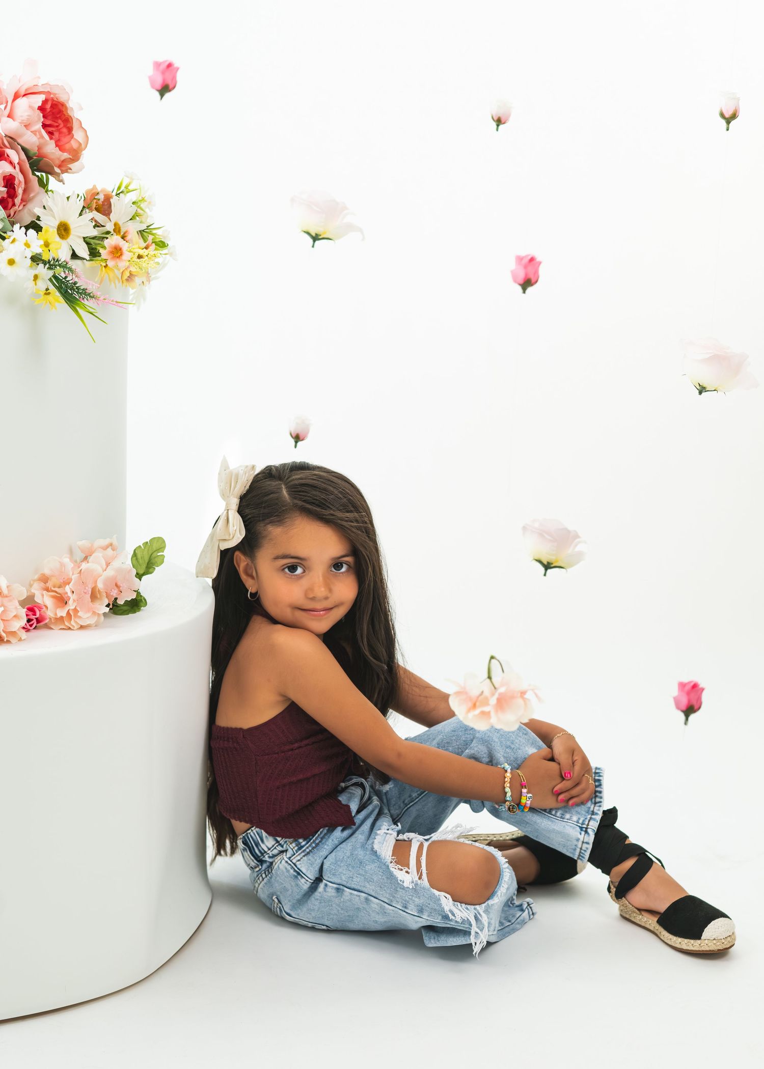 Young girl in ripped jeans and maroon top sits beside floral decorated white cylinder in photo studio.