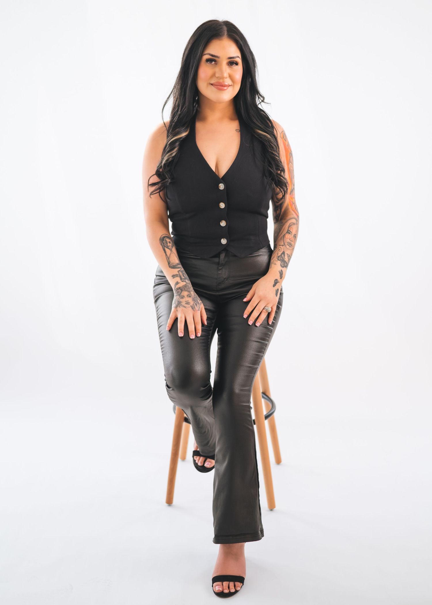 A person in a black vest and dark pants sits on a wooden stool against a white backdrop in a studio setting.