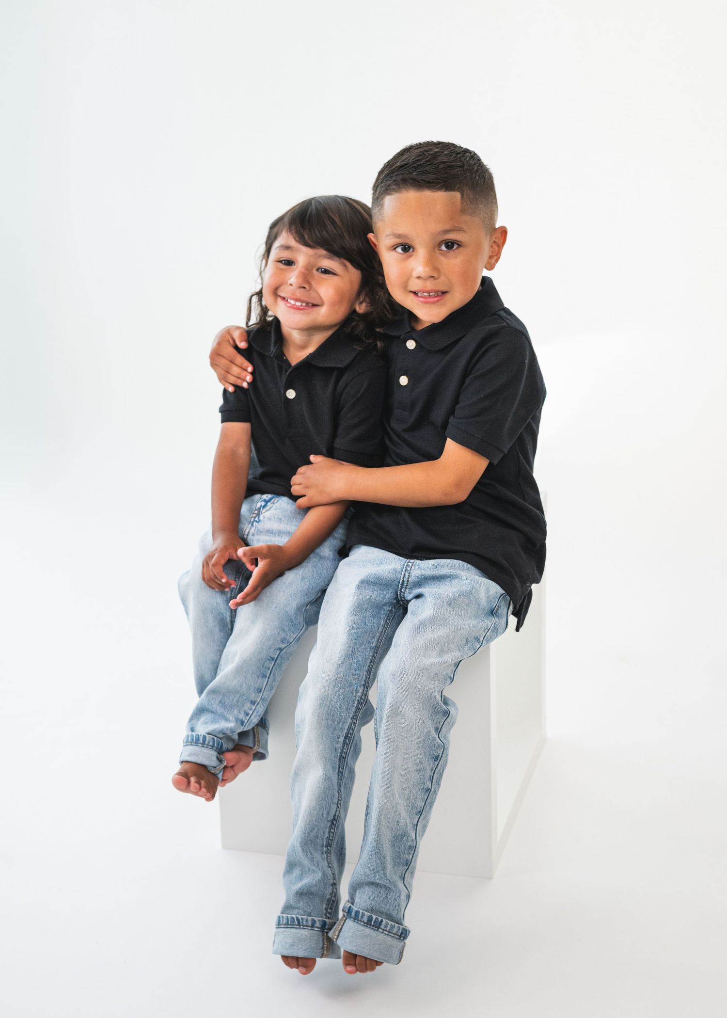 Two young siblings in matching black polo shirts and jeans sit together smiling on a white cube for a studio portrait.