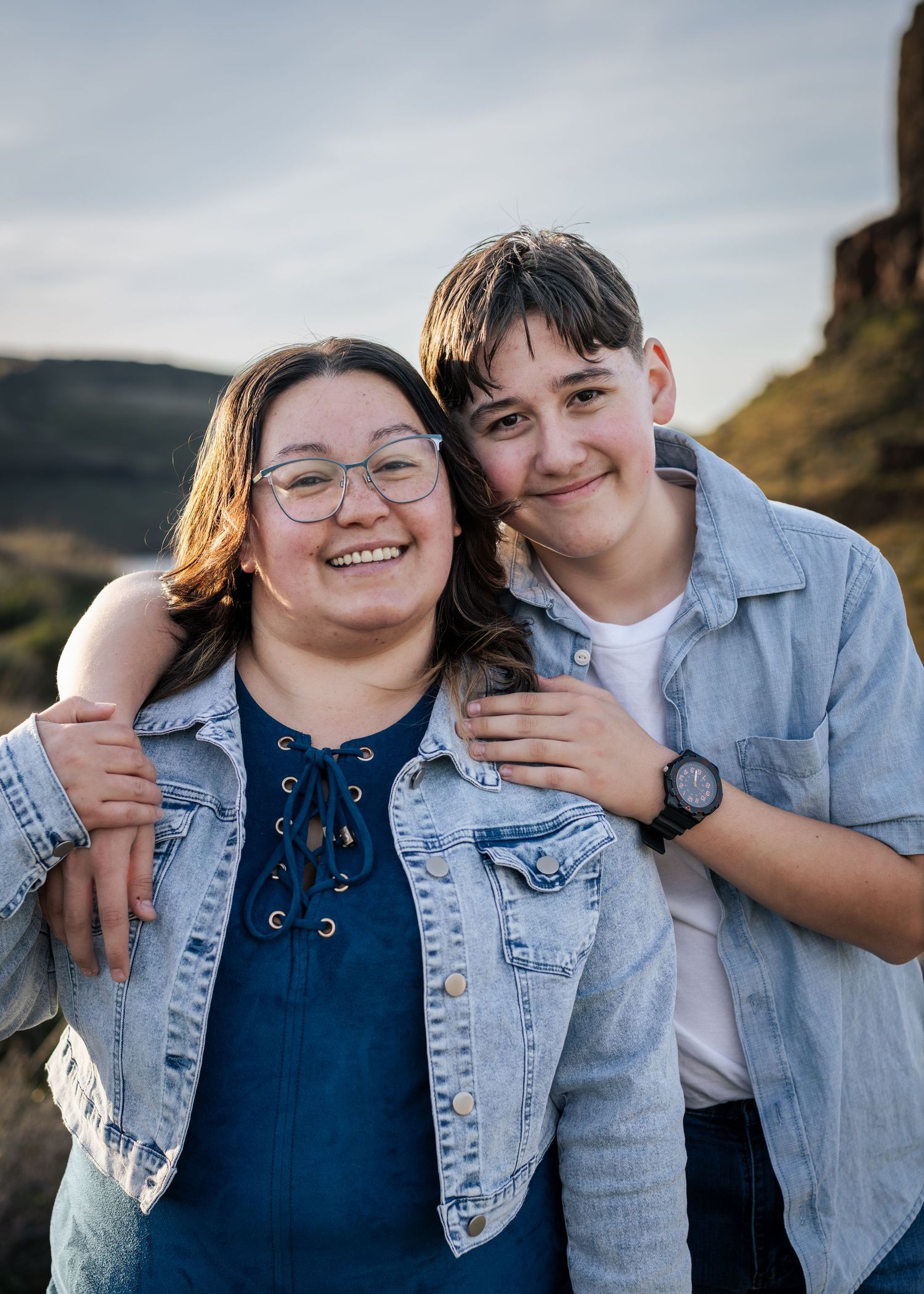 Two smiling young people in denim jackets pose together outdoors with rocky canyon landscape at golden hour.