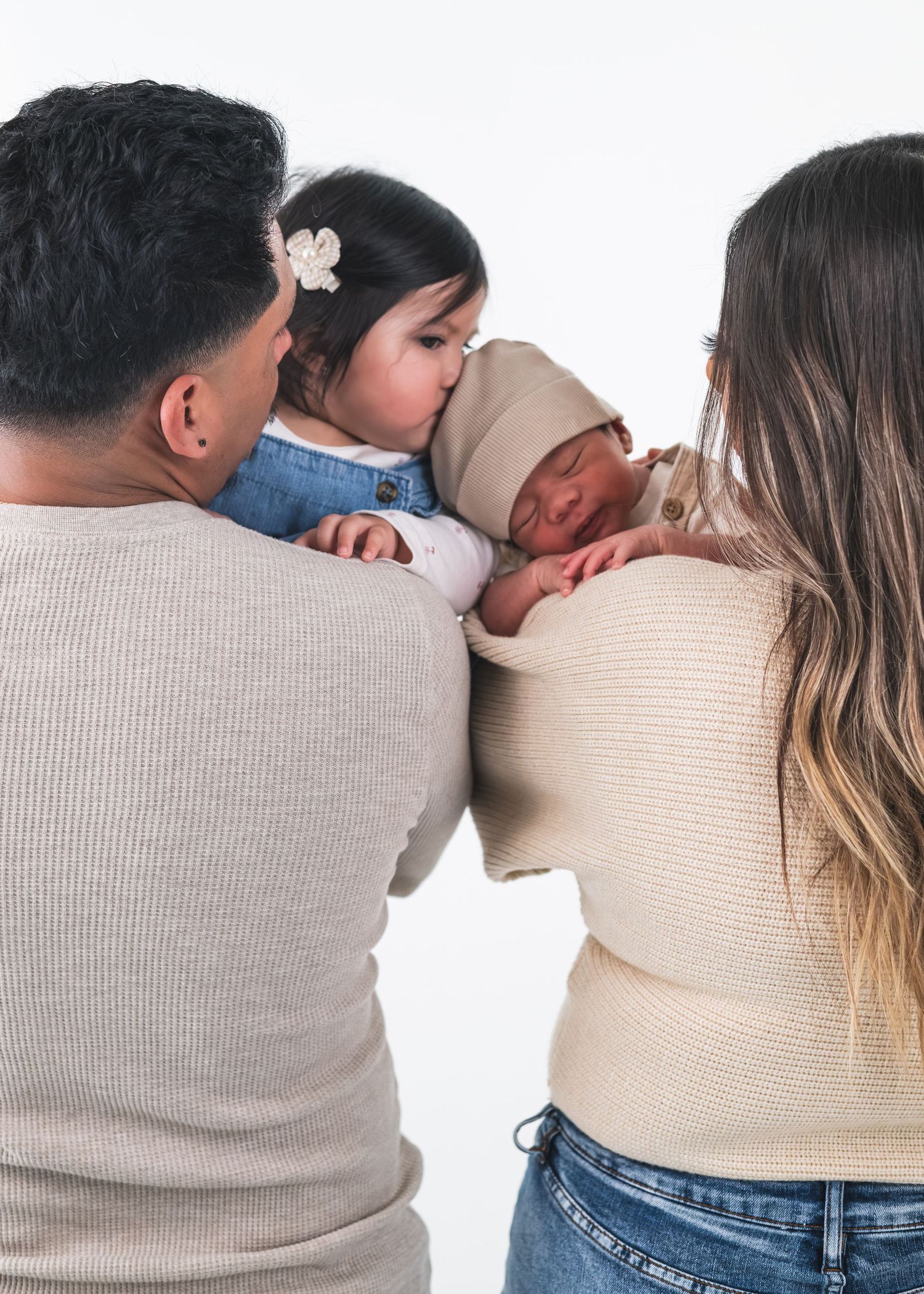 Parents holding a toddler girl kissing a newborn baby in a warm family portrait studio photo.