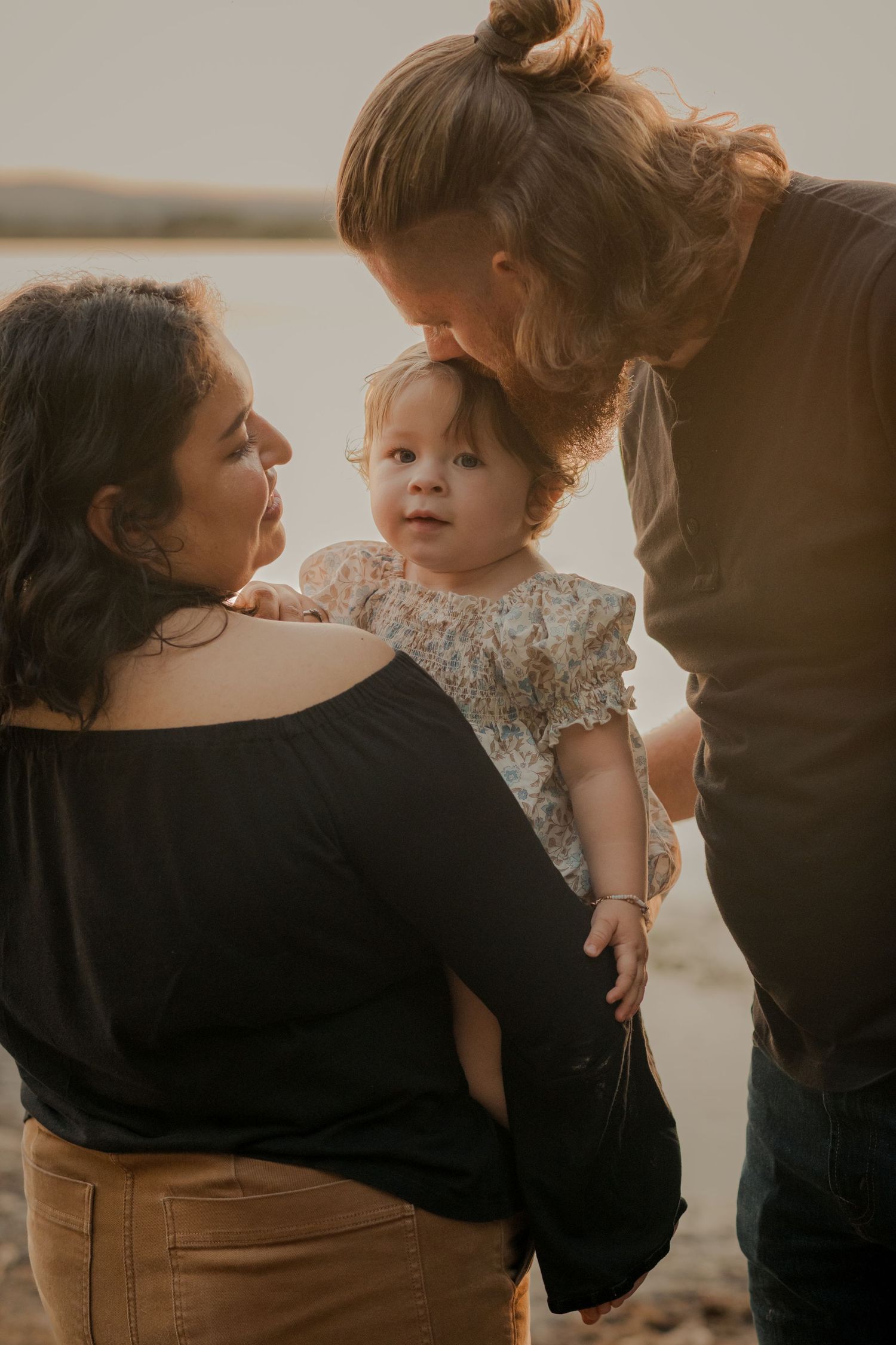 A tender family moment captured in warm lighting as parents embrace their young child wearing a floral dress.