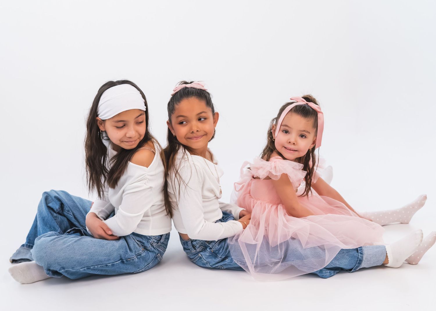 Three young girls sitting together on white background, wearing jeans and dresses, with hair accessories.