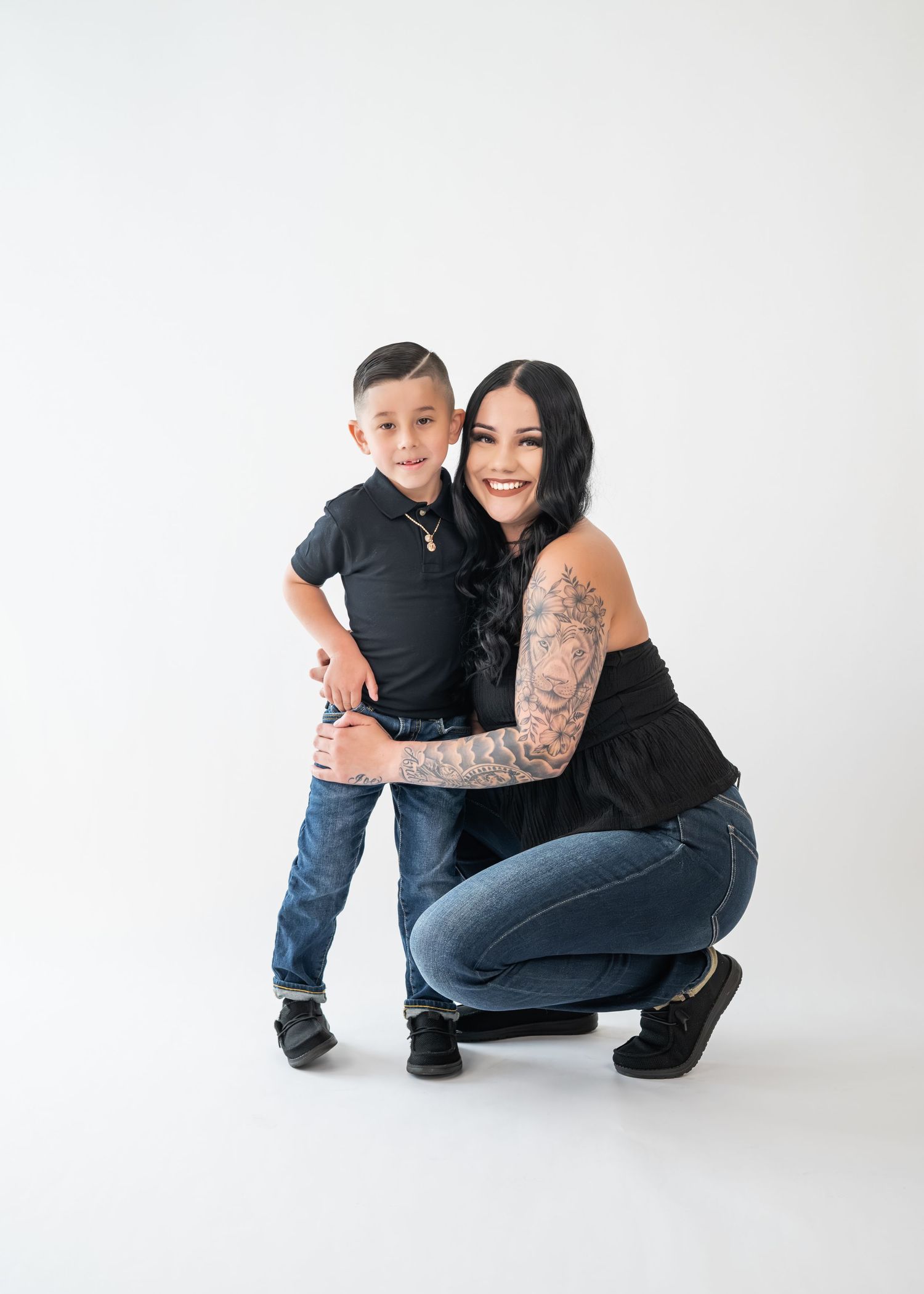 Mother with sleeve tattoos crouching beside young boy in black polo and jeans in white studio.