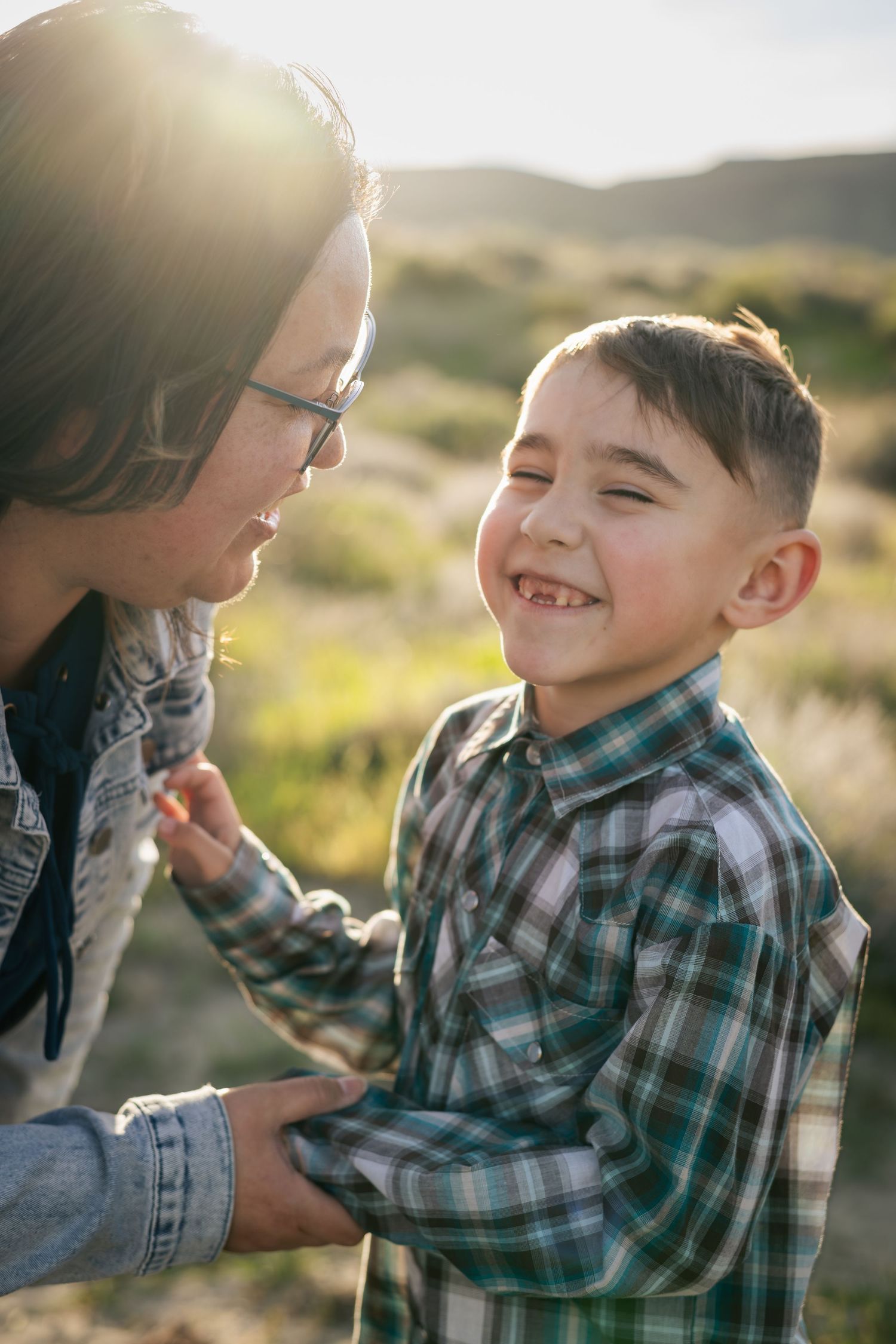 A joyful boy in a plaid shirt laughs with a woman wearing glasses outdoors in a sunny field.