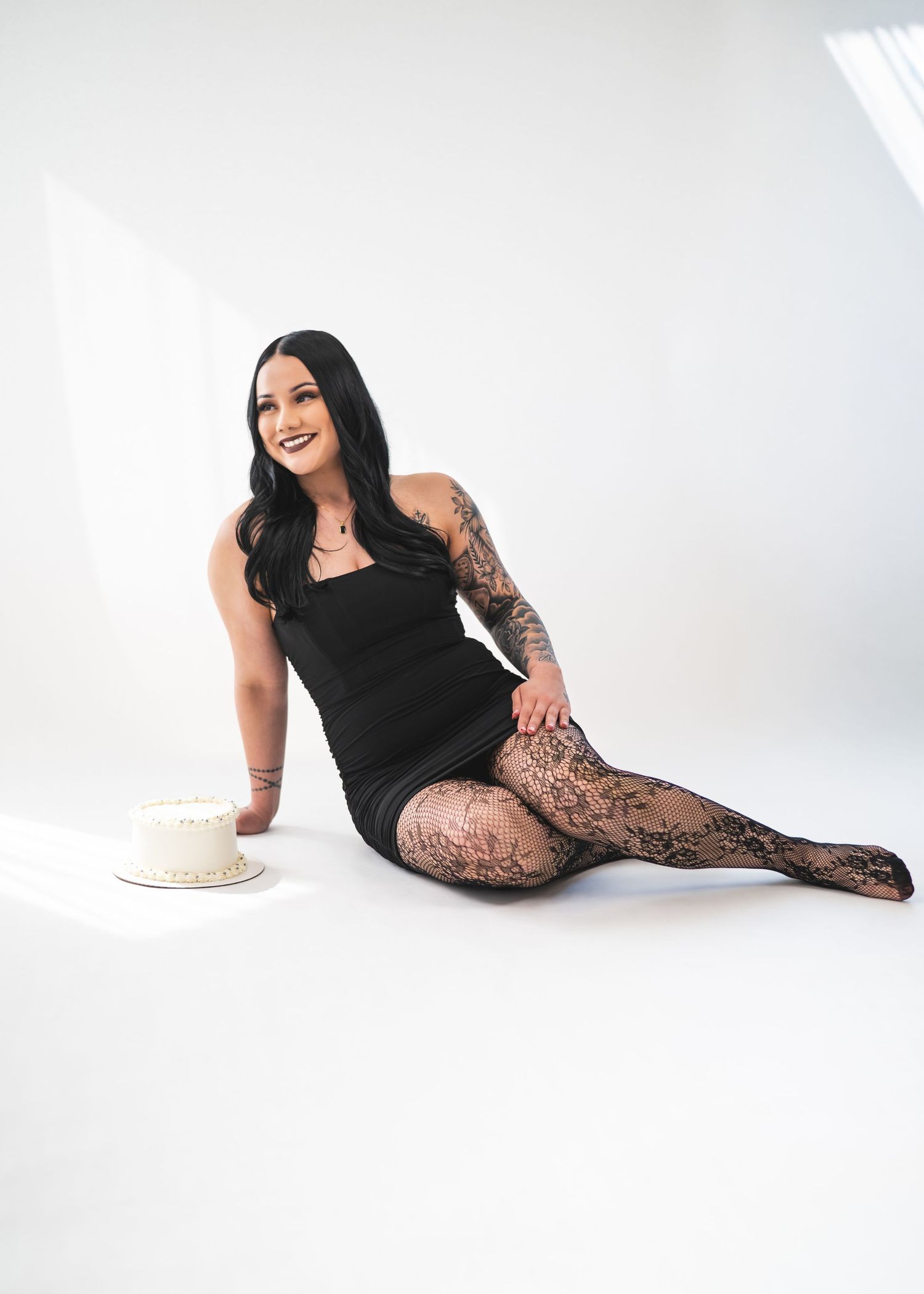 Woman in black mini dress and lace tights sits on white floor, smiling, with tattoos and a white cake nearby.