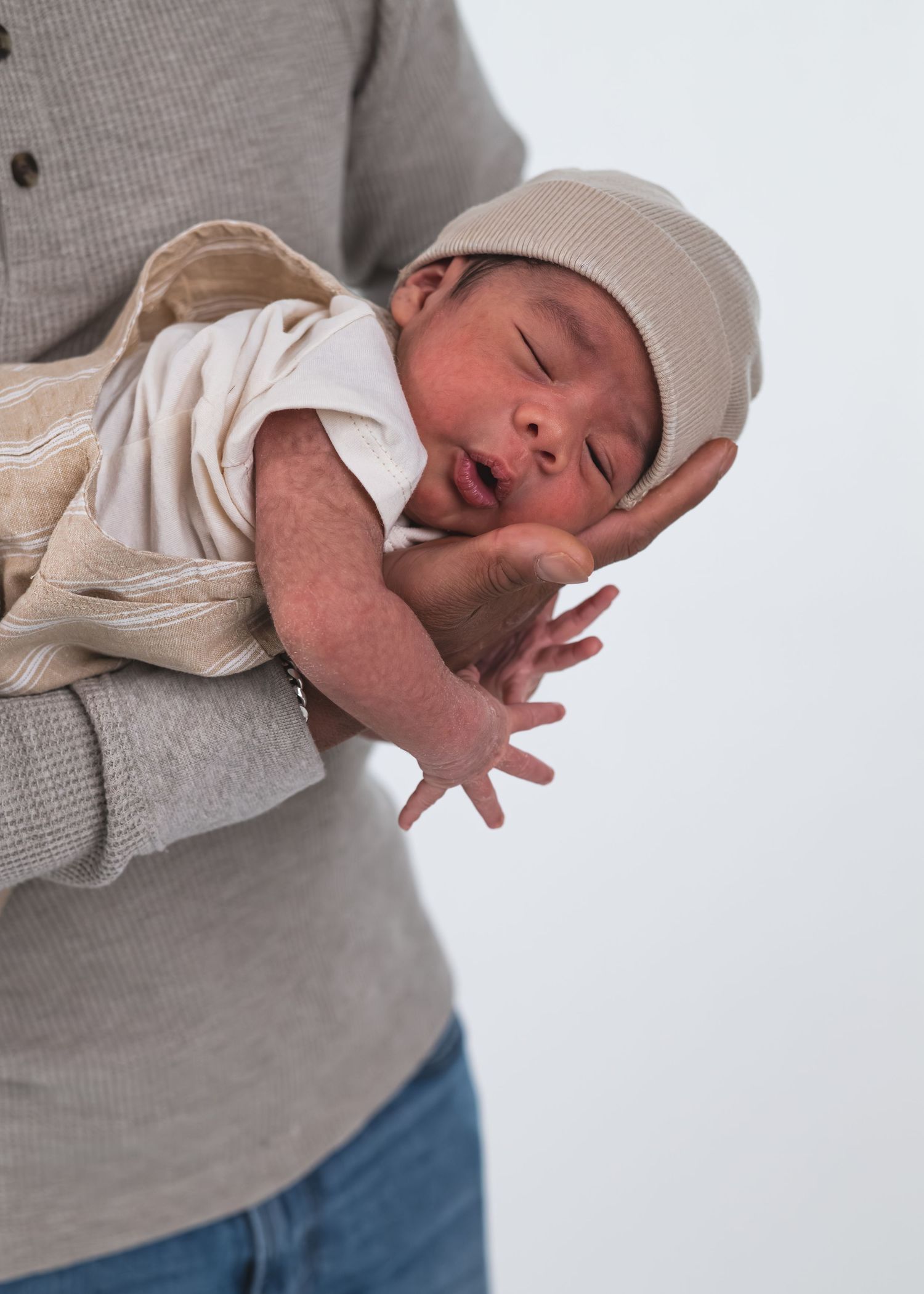 A parent cradles a sleeping newborn baby wearing a beige knit hat and white onesie against a white background.