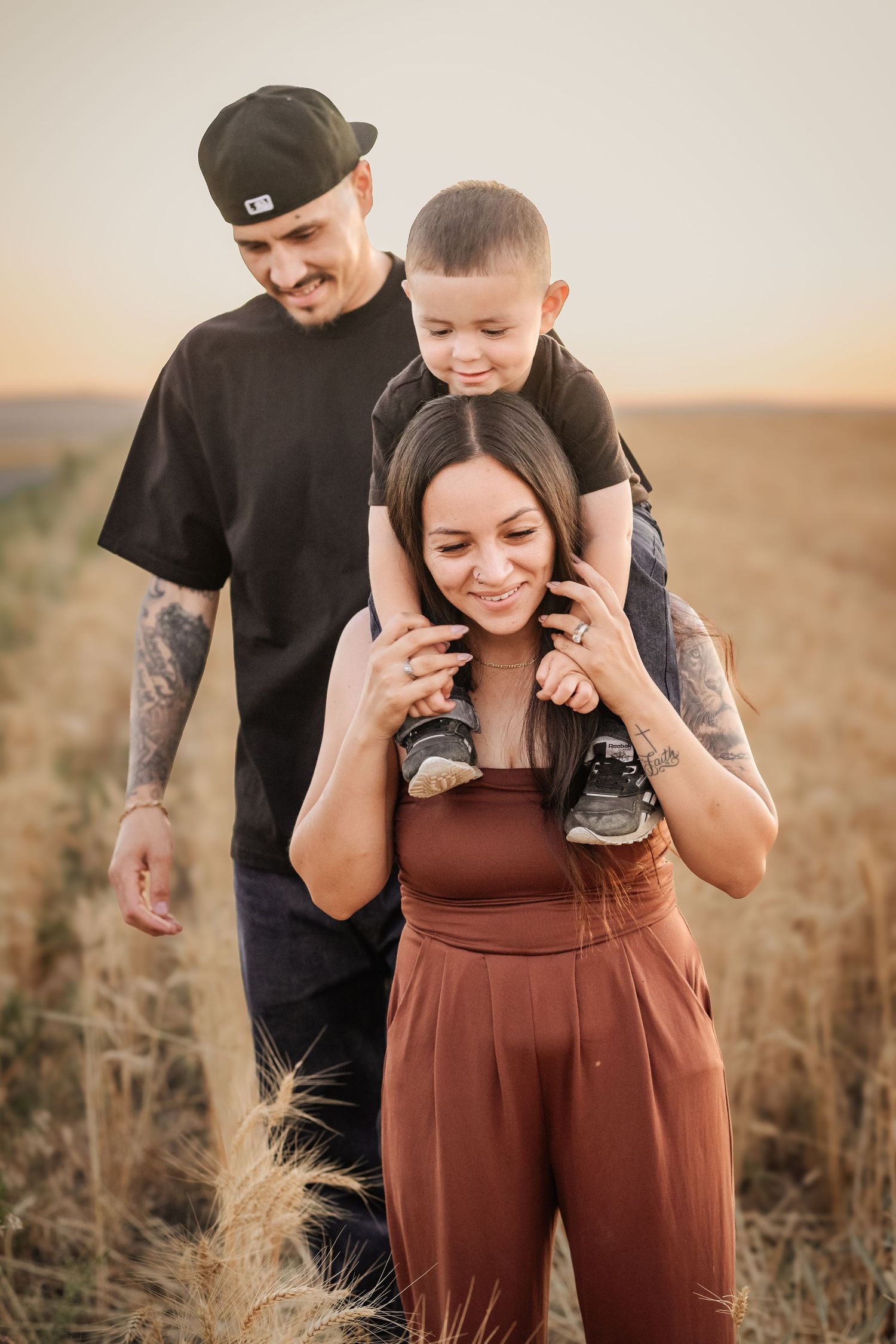 A happy family of three walks through a golden wheat field at sunset, with a young boy sitting on his mother's shoulders.