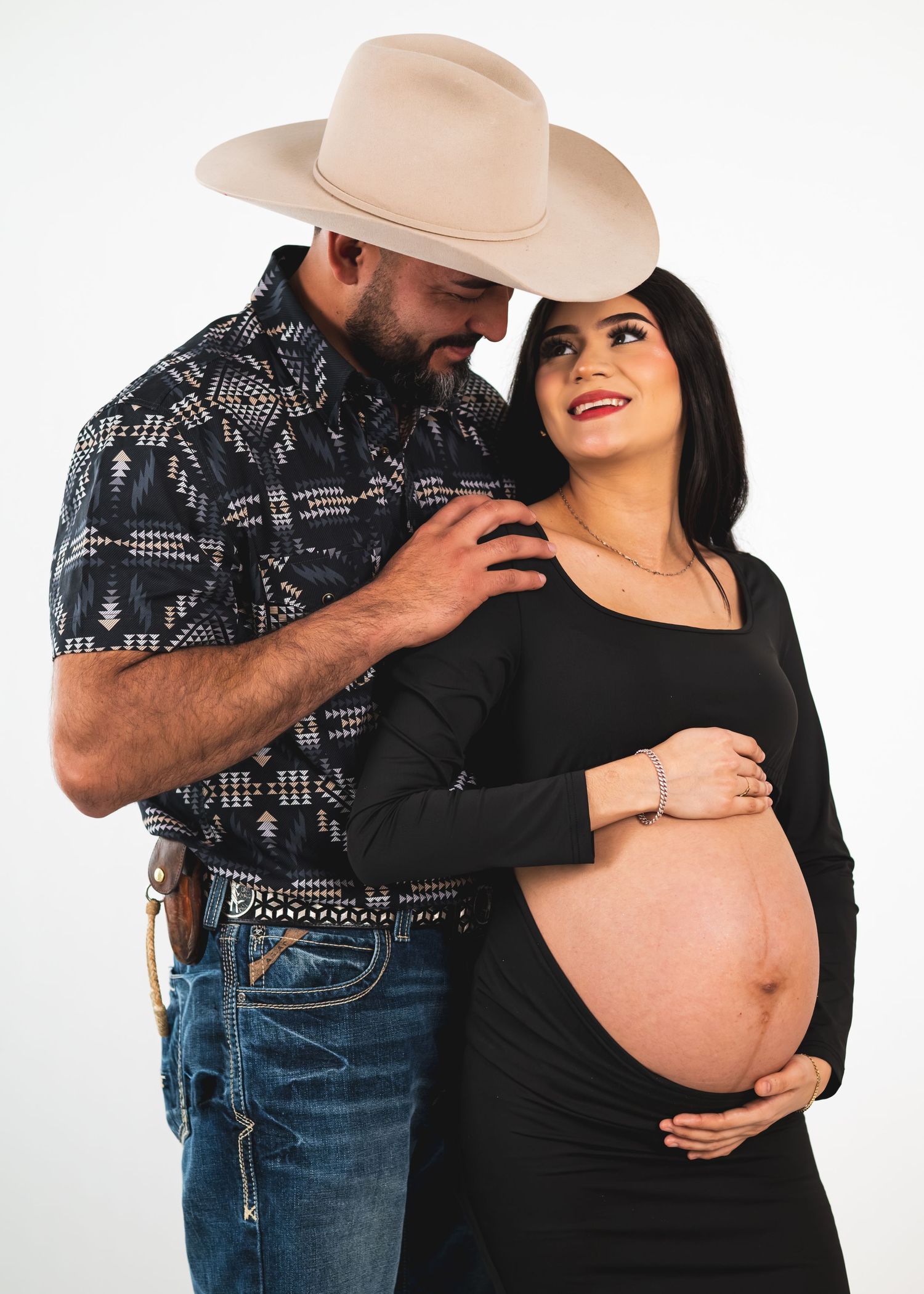 Couple in maternity photo shoot, man in cowboy hat and western shirt embraces pregnant woman in black dress.