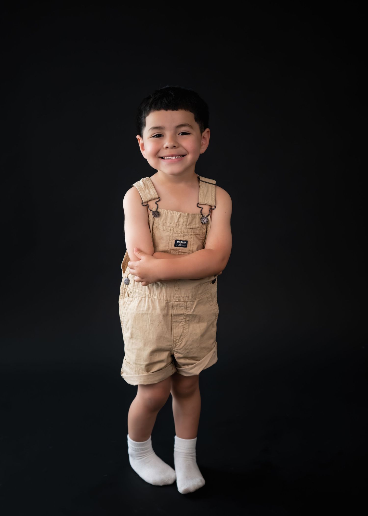 Smiling young boy in tan overalls and white socks poses with arms crossed against a dark studio background.