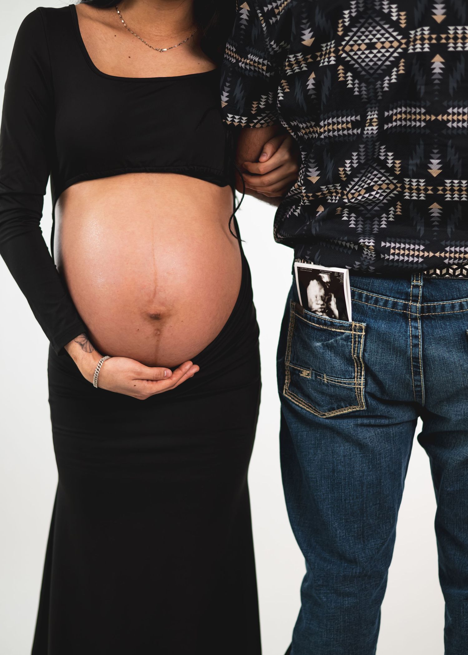 Pregnant couple maternity photo, woman in black crop top holding baby bump, man in jeans with ultrasound photo in pocket.