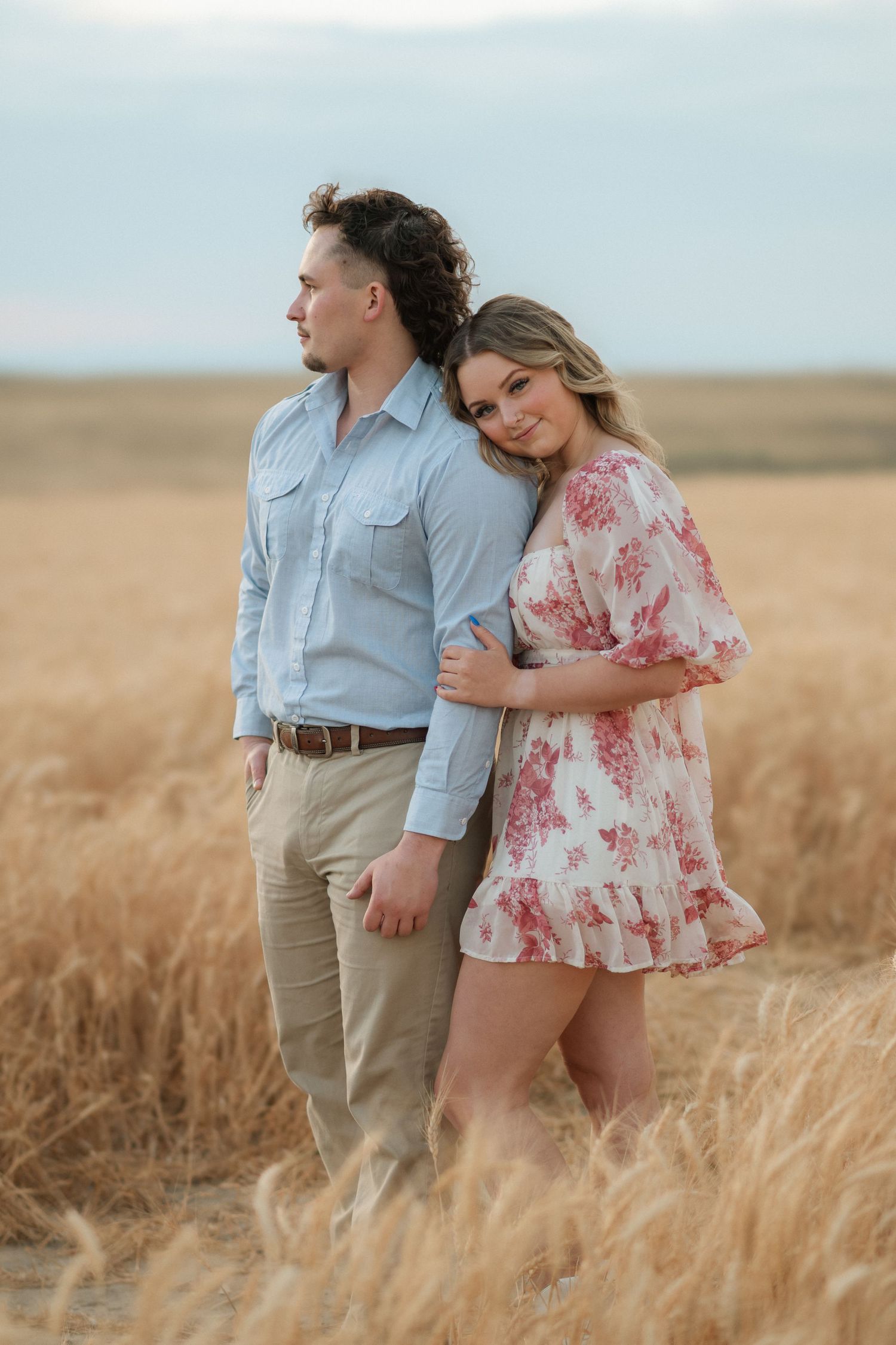 A couple embraces in a golden wheat field during sunset, with her wearing a floral dress and him in a blue shirt.