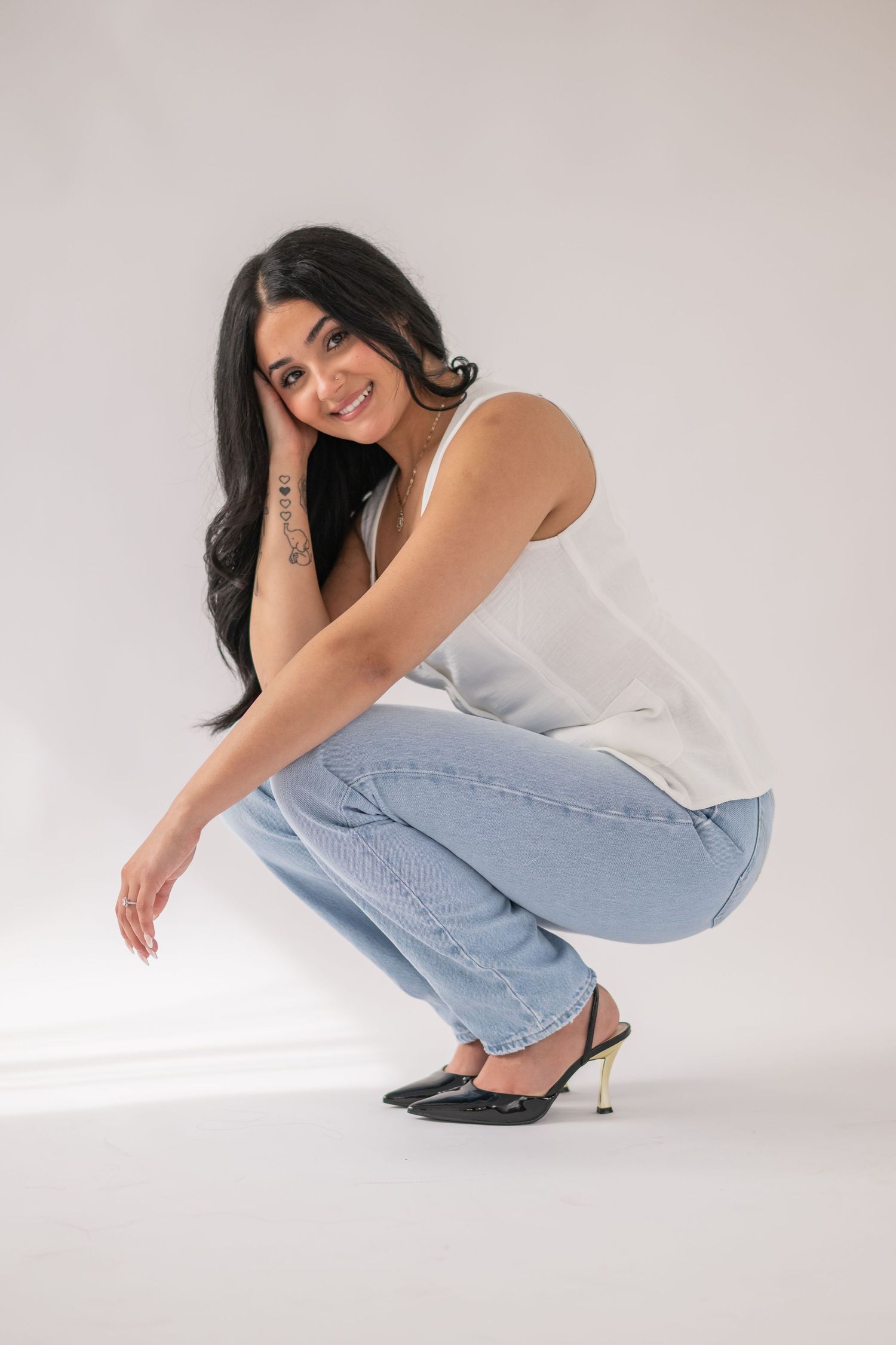 Person in casual white tank top and light blue jeans crouches while wearing black heels against white background.