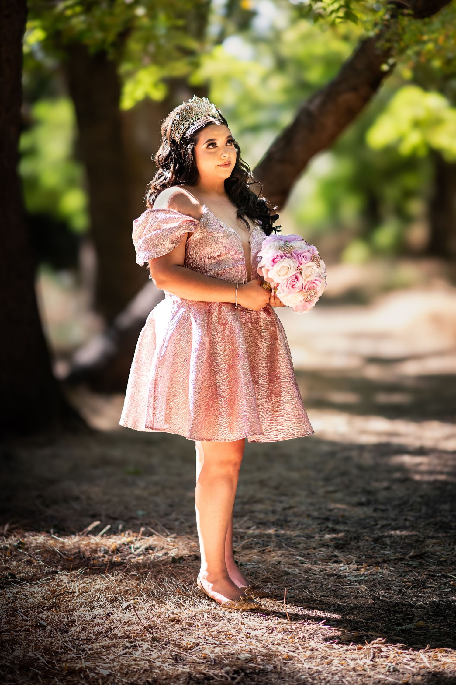 A small figure in a pink lace dress holds a bouquet while standing in dappled sunlight on a forest path.