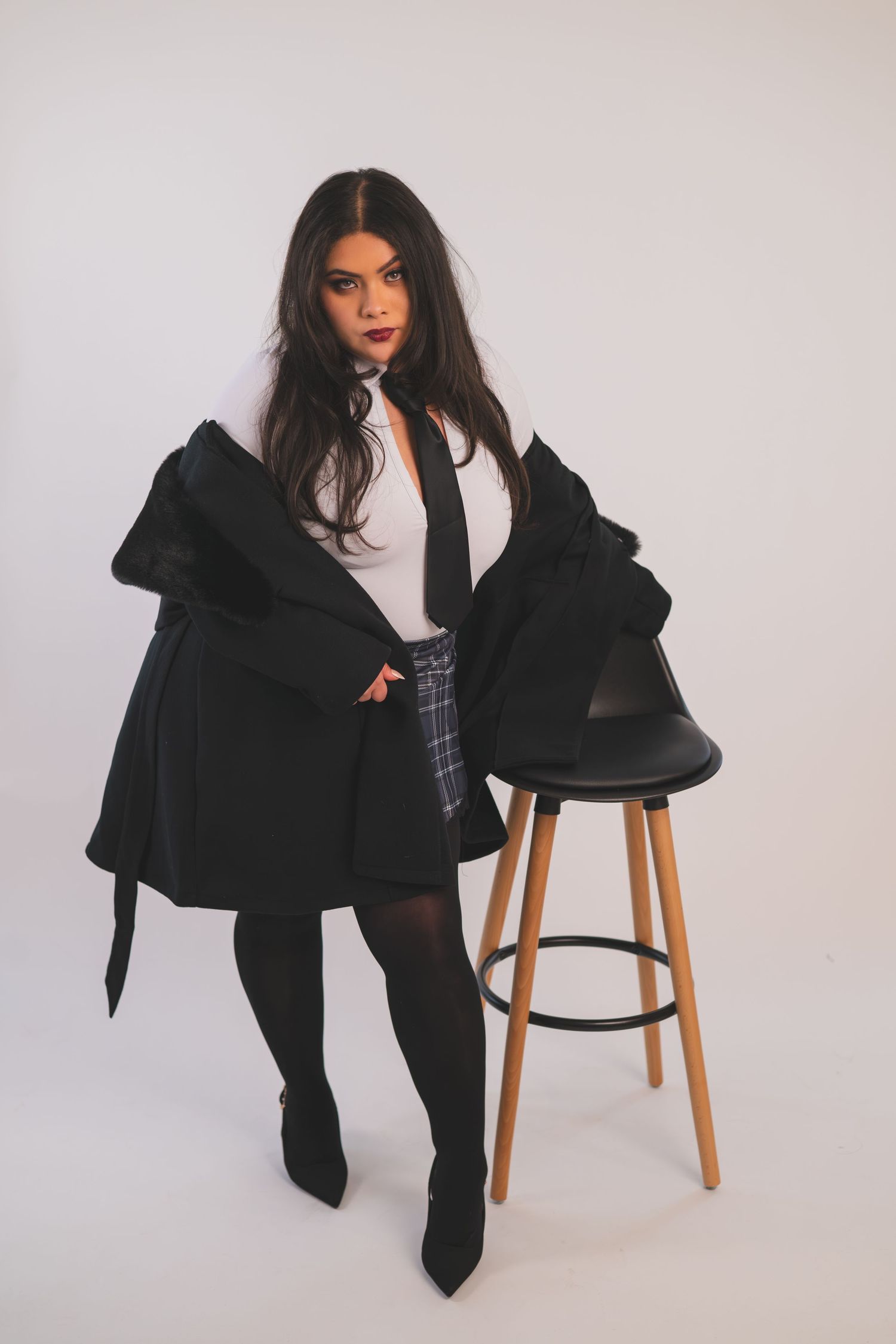 Woman in black coat, plaid skirt, and tights posing next to a bar stool against a white background.